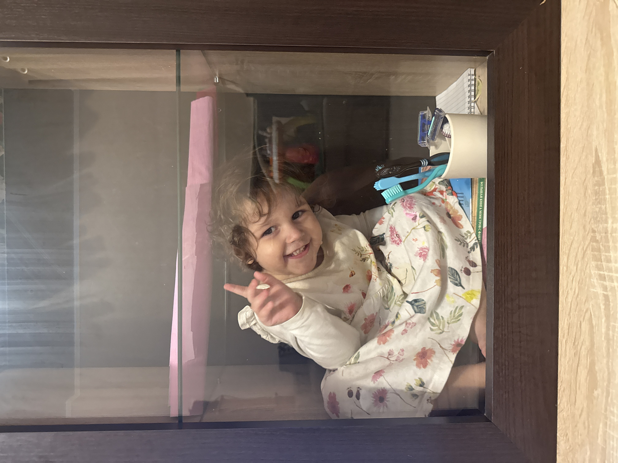 A young child with curly hair, wearing a light-colored floral dress and white long-sleeved shirt, is positioned inside what appears to be a glass-fronted display case or cabinet. The child is smiling and holding up one finger. Behind the child, within the cabinet, are various items, including books, toothbrushes, and a pink object. The cabinet has a dark brown wooden frame. The location is unidentifiable as Tobolsk, Russia from the image alone.