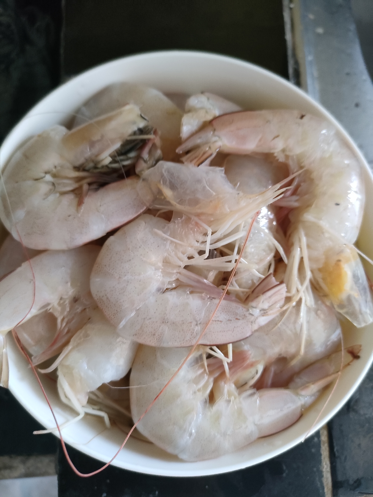 A close-up, overhead shot shows a white, oval-shaped bowl filled with raw, uncooked shrimp. The shrimp are a pale pinkish-white color and are arranged in a pile, with some overlapping. They appear to be whole, with heads and tails intact. Some of the shrimp have thin, red antennae or legs visible. The bowl is set on a dark, textured surface, possibly a countertop. The background is out of focus but appears to be a kitchen environment with some metallic elements, potentially part of a stove or sink. There are no people or discernible actions taking place. The lighting is neutral, suggesting an indoor setting. The location is Unknown, Indonesia.