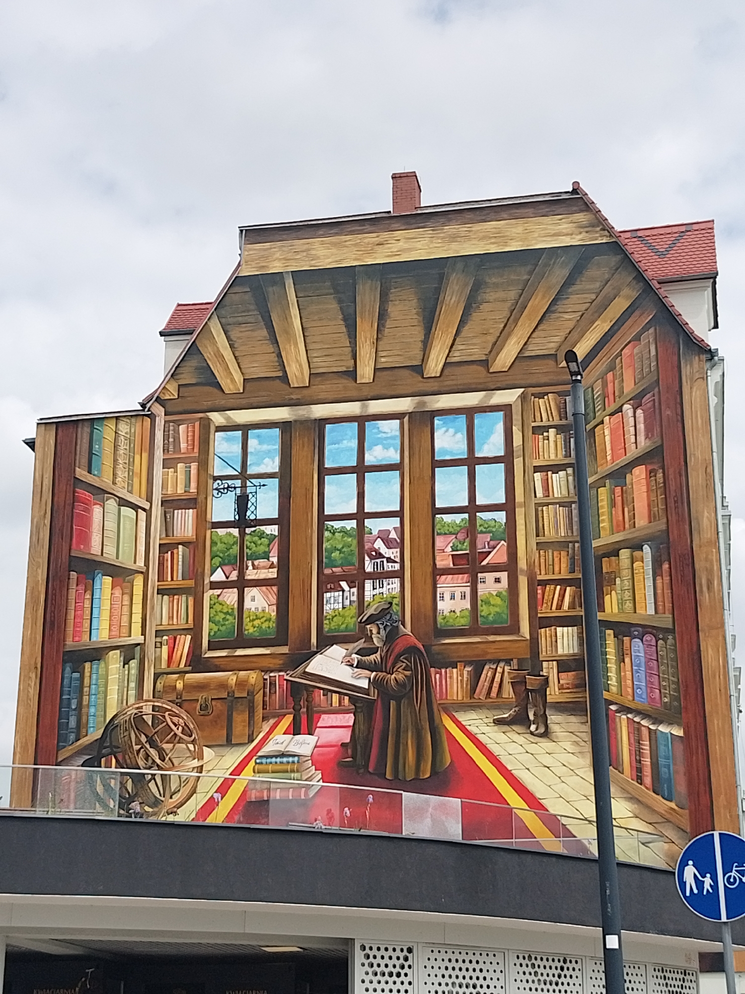 The image shows a building facade painted with an optical illusion. The artwork depicts a large library interior with wooden beams, bookshelves filled with books, a rug on a tiled floor, and a large window showing a cityscape. In the center of the painting, a man in a historical costume stands at a desk, writing. The overall impression is that the building is an extension of a large, inviting library. A pedestrian and bicycle sign is placed in the front of the building at the bottom right.