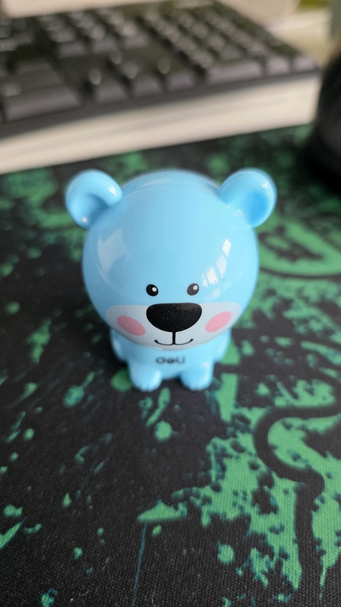 A close-up, eye-level shot shows a light blue, bear-shaped object with black eyes, a black nose, and pink cheeks. The object has a smiling mouth with the text "deli" printed below it. It is positioned on a dark green and black speckled surface, likely a mousepad. In the blurred background, a portion of a computer keyboard is visible. The image was taken indoors, suggesting a home or office environment. There are no people present, and no specific location in Brest, Belarus, can be identified from the visual information.