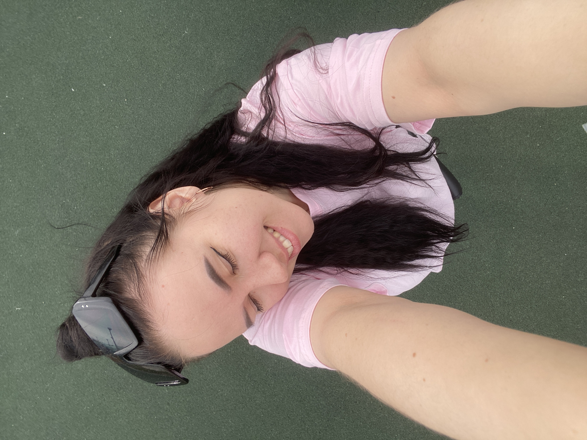 A woman with long, dark hair and sunglasses perched on her head takes a selfie outdoors. She wears a light pink t-shirt. She is smiling slightly with her eyes mostly closed. The background is a solid, dark green surface. This is a close-up shot. The location, Perm, Russia, cannot be definitively determined from the image alone.