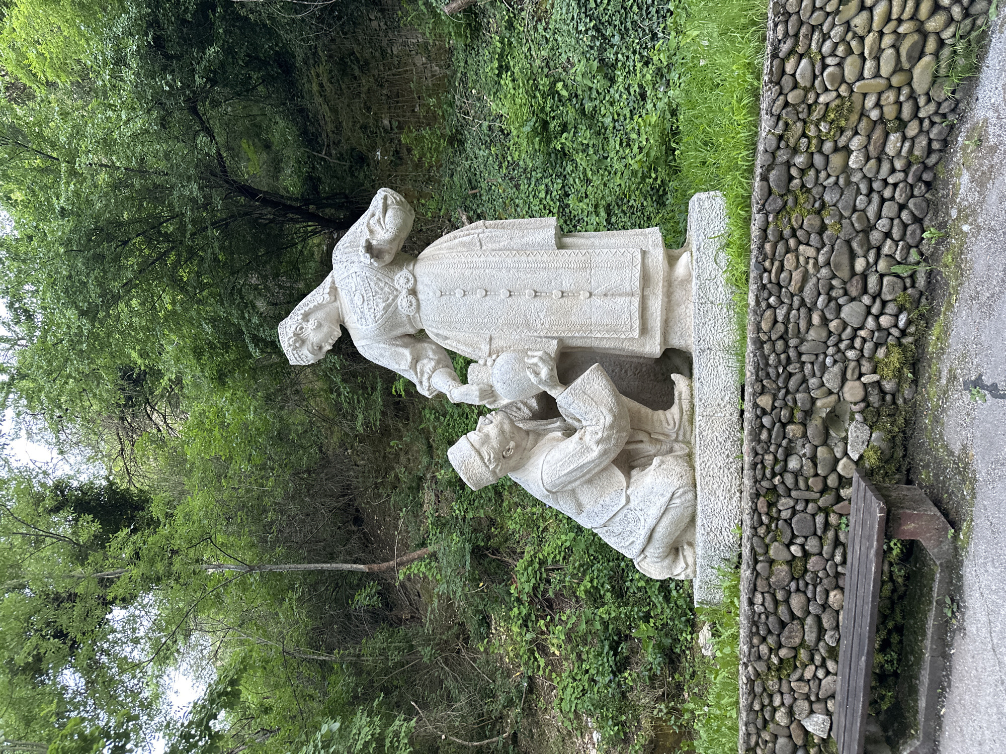 The image depicts a light-colored stone sculpture of two figures. One is a standing woman in traditional clothing, likely Bulgarian folk costume, placing her hands on a kneeling man who is holding a bowl. The sculpture is situated outdoors, next to a rock wall overgrown with greenery and grass. Behind the sculpture, there is a lush green backdrop of trees and foliage. The specific location is in the city of Lovech, Bulgaria.