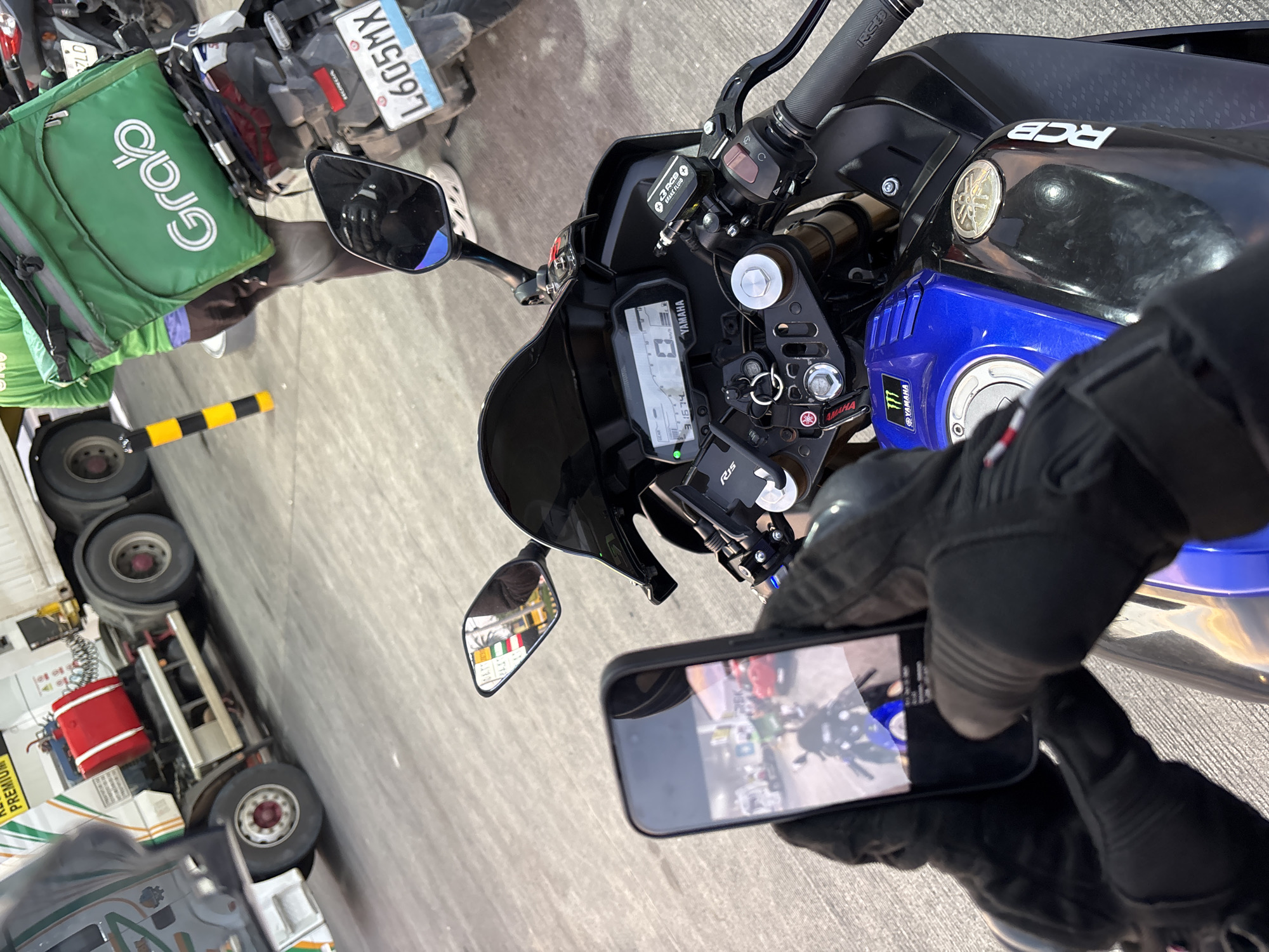 The image depicts a close-up view of a motorcycle, likely a Yamaha R15, parked at a gas station in Davao City, Philippines. The rider, wearing black motorcycle gloves, is holding a smartphone in their left hand, capturing a photo of the motorcycle's dashboard and a portion of the gas station environment.
In the foreground, the motorcycle's handlebars, instrument cluster displaying "0" km/h and "31674" kilometers, and part of the fuel tank are visible. The fuel tank is predominantly blue and black, with a metallic fuel cap and "RCB" branding. A prominent "Yamaha" logo with a Monster Energy emblem is also visible on the blue section of the tank.
The background reveals a large white truck with a red cylindrical tank, suggesting it is a fuel tanker. A yellow and black striped bollard is positioned near the truck. To the right of the truck, another motorcycle, possibly a Grab delivery motorcycle given the green Grab delivery bag attached to its rear, is parked. The license plate on this motorcycle is visible and reads "L605MX".
The ground appears to be paved with concrete. The overall lighting suggests it is daytime. The presence of the truck, gas pumps (implied by the gas station setting), and the delivery motorcycle strongly indicate an urban environment within Davao City, likely a well-trafficked area due to the presence of commercial vehicles and delivery services.