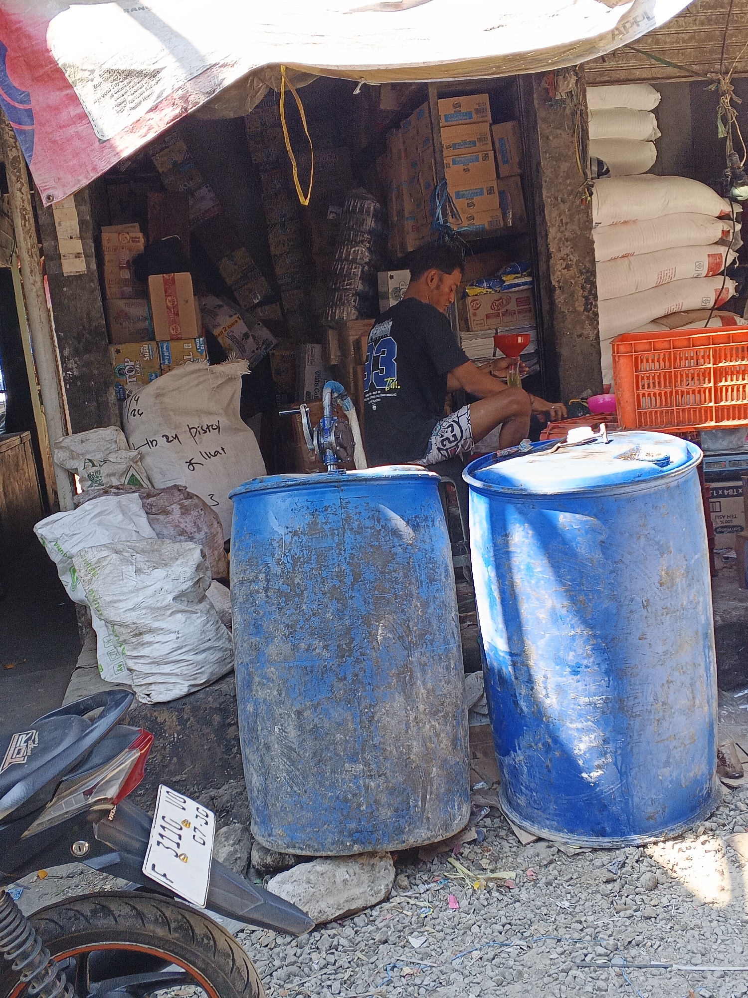 The image depicts an outdoor market stall in Warungkiara, Indonesia, likely selling various goods. In the foreground, two large blue plastic barrels are prominent, one on the left and one on the right. To the left of the barrels, a black motorcycle is parked, with a white license plate displaying "F 3116 VO 07-30". Behind the barrels, a man in a black t-shirt and patterned shorts is seated, appearing to be engaged in pouring liquid from a funnel into a small container, possibly for sale or use. The stall is filled with stacked boxes and large white sacks, suggesting a variety of inventory. The environment appears somewhat cluttered and rustic, typical of a local market setting. The ground is covered with gravel and debris, and overhead, a tarp or awning provides shade.