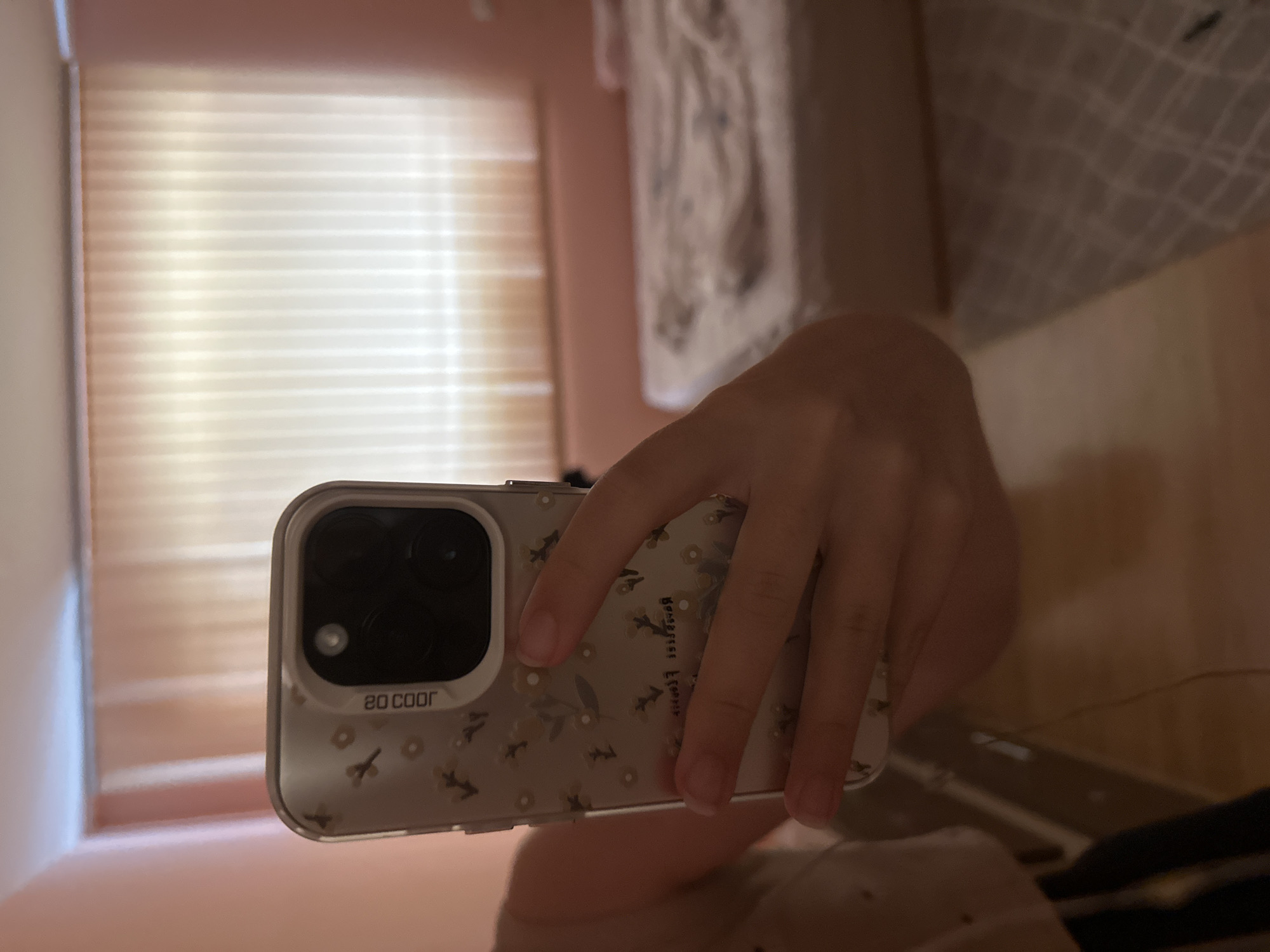 The image shows a person's hand holding a smartphone in a mirrored reflection. The hand is in the foreground, holding a light-colored phone case with a floral pattern. The phone's camera bump is visible on the back of the case. In the reflection, we can see a window with horizontal blinds, suggesting an indoor setting. There is also a hint of what appears to be a white fabric with a pattern, possibly a curtain or clothing, hanging in the background of the reflection. The lighting appears to be natural, possibly from the window. The overall impression is a casual, everyday moment captured through a reflection. Visible text on the phone case in the reflection reads "20 COOR" backwards.