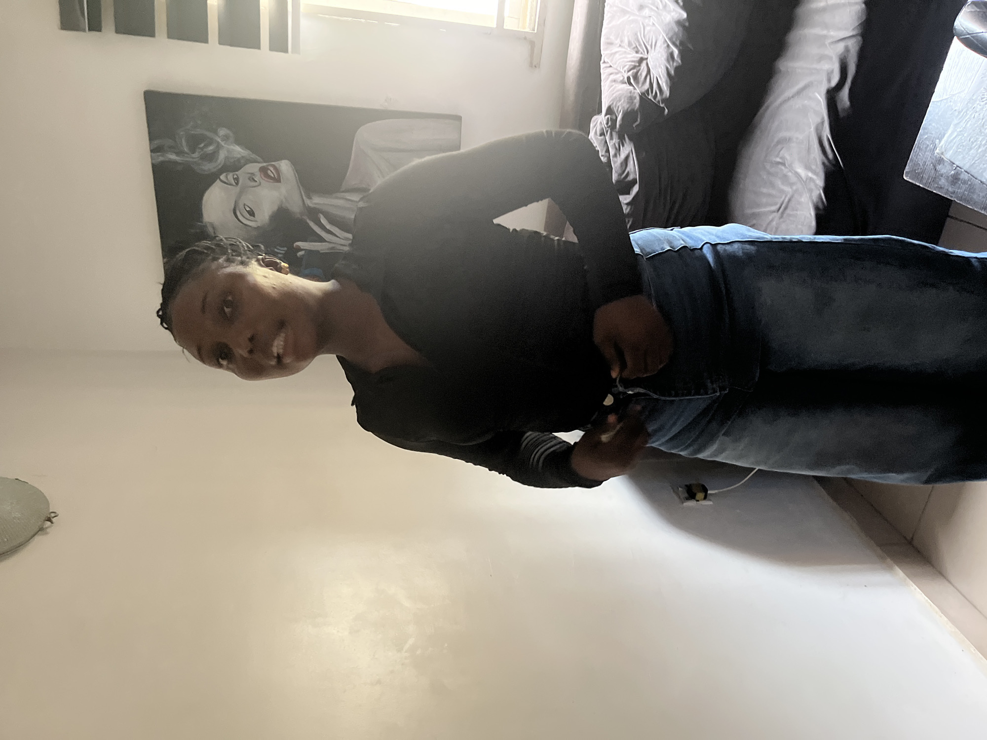 A young Black woman stands in what appears to be a bedroom in Abuja, Nigeria. She is wearing a long-sleeved black top and a denim skirt. Her hair is styled in braids. She is looking towards the camera and adjusting her skirt. In the background, there is a painting of a woman with smoke around her face, a window with blinds, and a bed with dark linens. The walls are painted a light color, and the floor is tiled. An electrical outlet is visible on the wall near the floor.