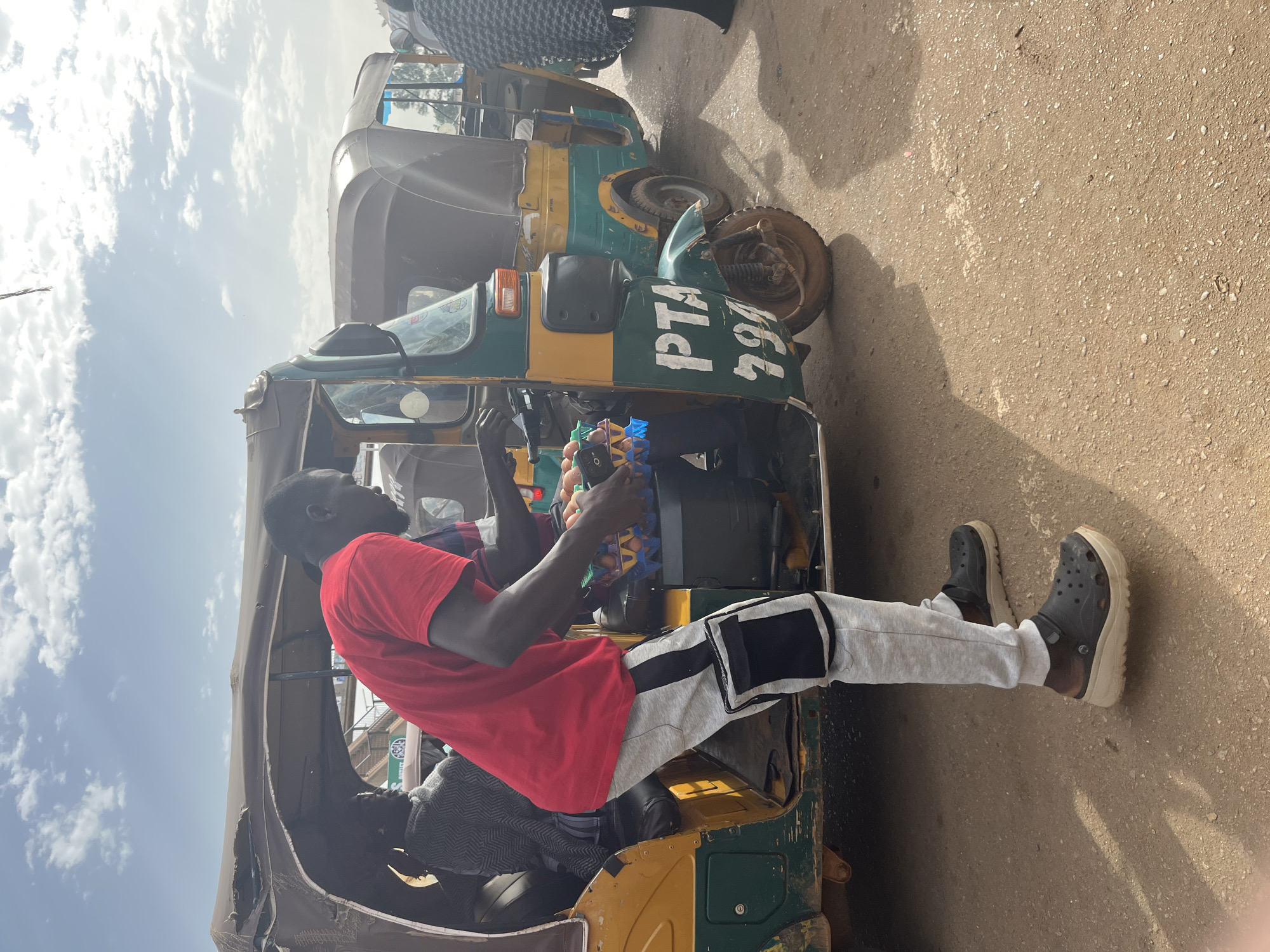 An overhead shot captures a man in a red t-shirt and gray sweatpants with black accents, wearing black crocs, standing next to a teal and yellow auto-rickshaw. He holds a crate of eggs in his hands. Two other individuals are visible inside the auto-rickshaw, one wearing a gray patterned sweater and the other mostly obscured. The auto-rickshaw has "PTA 294" painted on its side. The ground appears to be dirt or unpaved road. The scene suggests a busy marketplace or transportation hub in Gura Top, Nigeria, under a bright, partly cloudy sky.