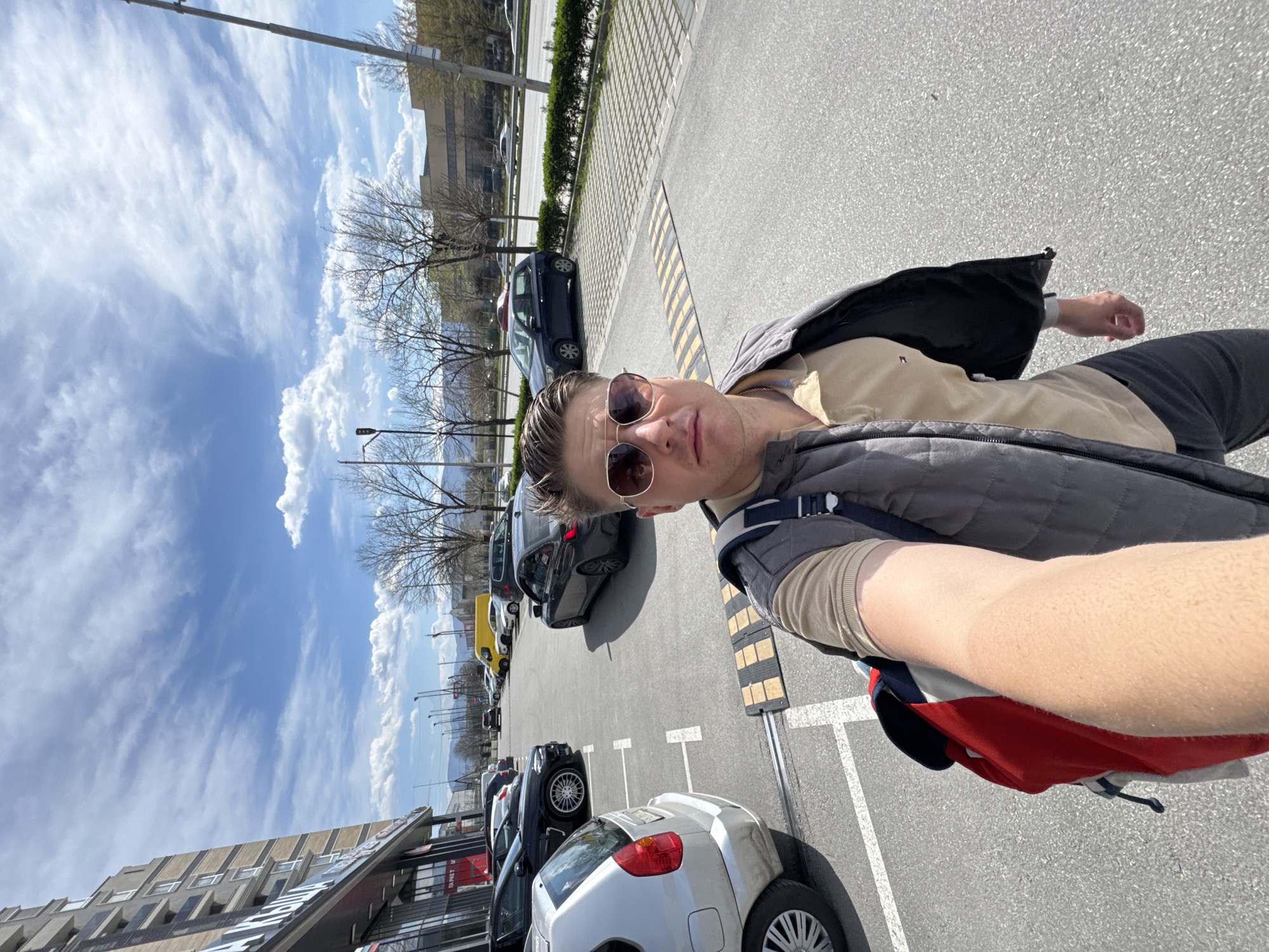 A man is taking a selfie in a parking lot in Plovdiv, Bulgaria. He is wearing sunglasses, a beige shirt, a grey vest, and a backpack. The parking lot is full of cars, and there are trees and buildings in the background. The sky is blue with clouds.