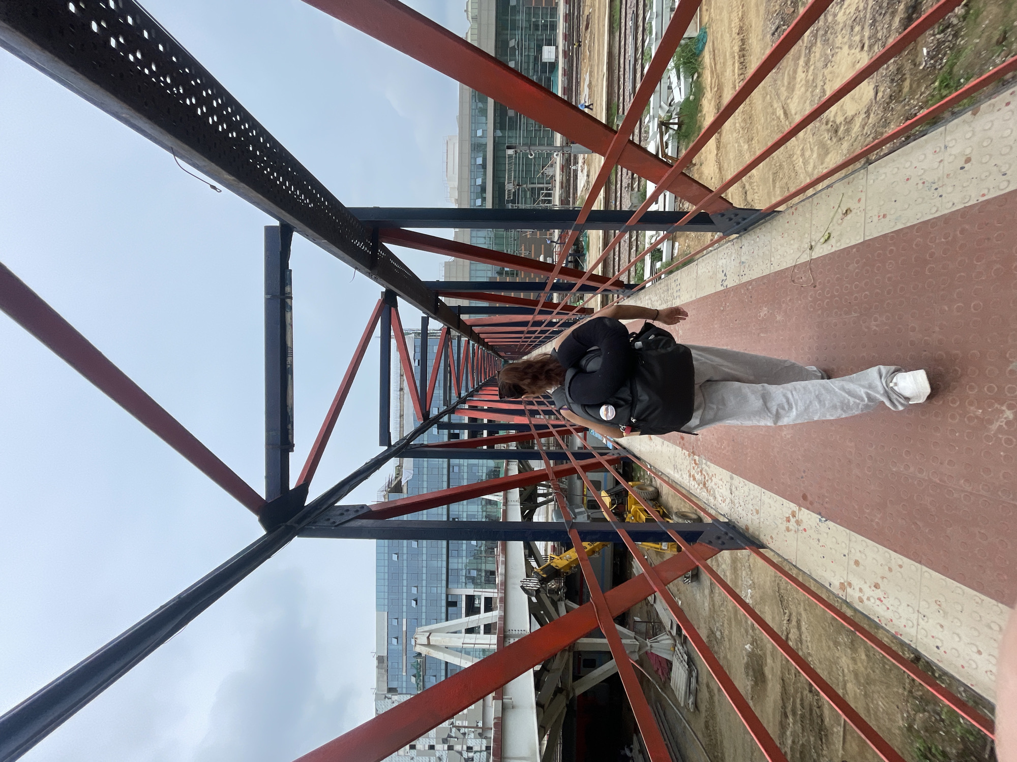 The image depicts a person with a backpack walking on a red and white footbridge. The footbridge has red metal railings and a dark metal frame. In the background, there is a modern building with glass windows, and construction activities are visible, including a yellow crane. The scene appears to be in New Delhi, India, possibly near a transportation hub or a construction site. The sky is overcast.