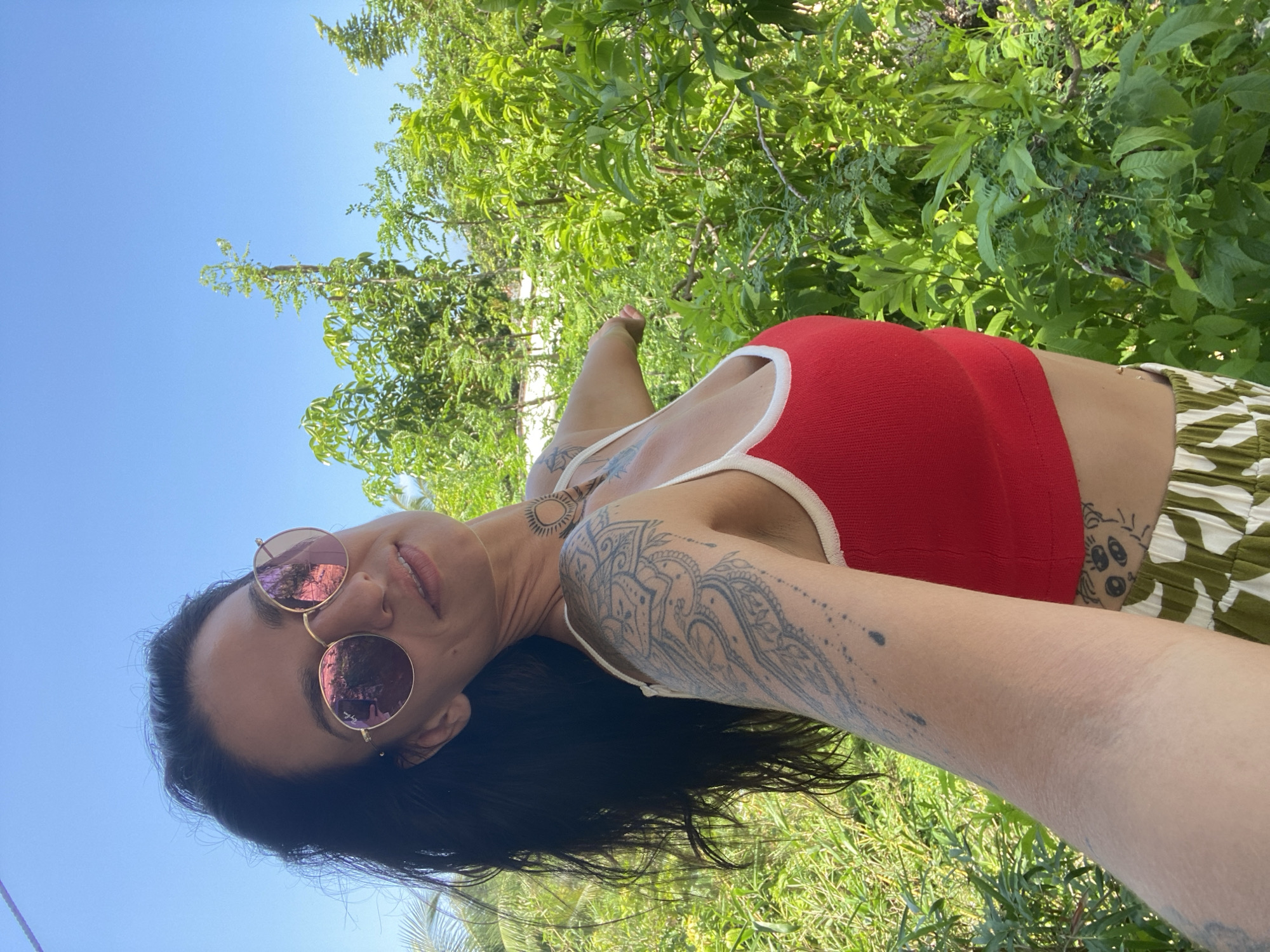 A woman with long, dark hair and tattoos on her arm and shoulder is taking a selfie outdoors. She is wearing round, rose-tinted sunglasses, a red tank top with a white trim, and patterned pants. She is standing in front of lush green foliage, with a clear blue sky above. The scene appears to be in a tropical or sub-tropical location, potentially Mũi Né, Vietnam, known for its beaches and coastal scenery.