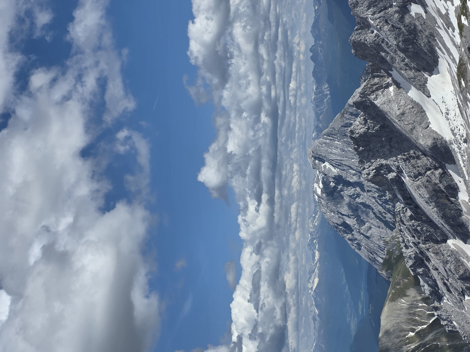 The image depicts a dramatic mountain landscape, likely in the Bavarian Alps near Garmisch-Partenkirchen, Germany, given the characteristic jagged peaks and snow-dusted slopes. In the foreground, rugged, grey rocky mountain ridges dominate, interspersed with patches of lingering snow. These ridges descend sharply, hinting at a steep, high-altitude environment. Further back, a prominent, craggy mountain peak rises, its summit capped with snow and its slopes etched with rock. Beyond this central peak, a vast expanse of layered mountain ranges recedes into the distance, with the furthest peaks appearing as soft, blue silhouettes against the horizon. A hazy valley or lower terrain is visible between the closer and further mountain ranges. The sky above is a vibrant blue, populated by a dramatic array of white and grey clouds. The clouds are thick and textured, with some appearing more wispy and others dense and cumulus-like, casting shadows that enhance the mountainous terrain's dramatic contours. A faint white contrail is visible in the upper right quadrant of the sky, indicating the presence of an aircraft high above. The scene conveys a sense of grandeur, wildness, and the immense scale of the natural environment. There are no people or discernible human activity in the frame.