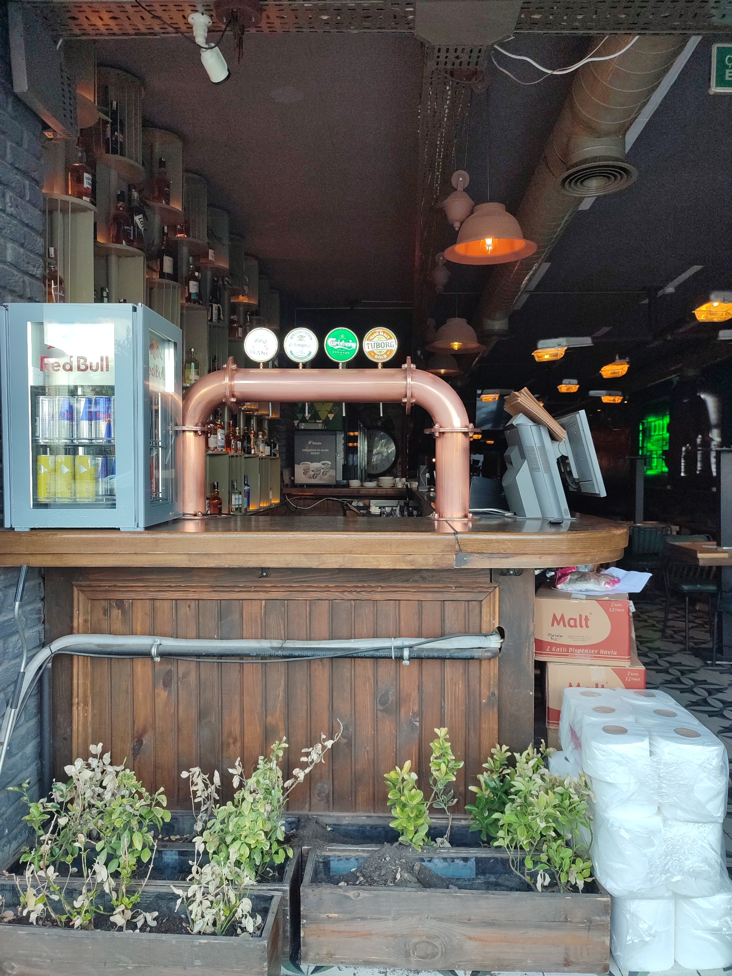 The image captures a street-level view of a bar or pub entrance in Kadıköy, Turkey. In the foreground, two wooden planters contain wilting plants. To the right, stacked cardboard boxes labeled "Malt" and packages of toilet paper are visible. The bar itself features a wooden counter and a copper beer tap system with four dispensers, each displaying a different beer logo: "1904 BLANC," "La Trappe," "Carlsberg DENMARK," and "TUBORG GOLD." A small, illuminated Red Bull mini-fridge sits to the left of the bar, stocked with cans. Behind the bar, shelves stocked with bottles are dimly lit, and a cash register system with two monitors is present. The ceiling is adorned with industrial-style lighting fixtures and exposed ductwork. The overall ambiance suggests a casual establishment, potentially a pub or a bar, on a street in Kadıköy.