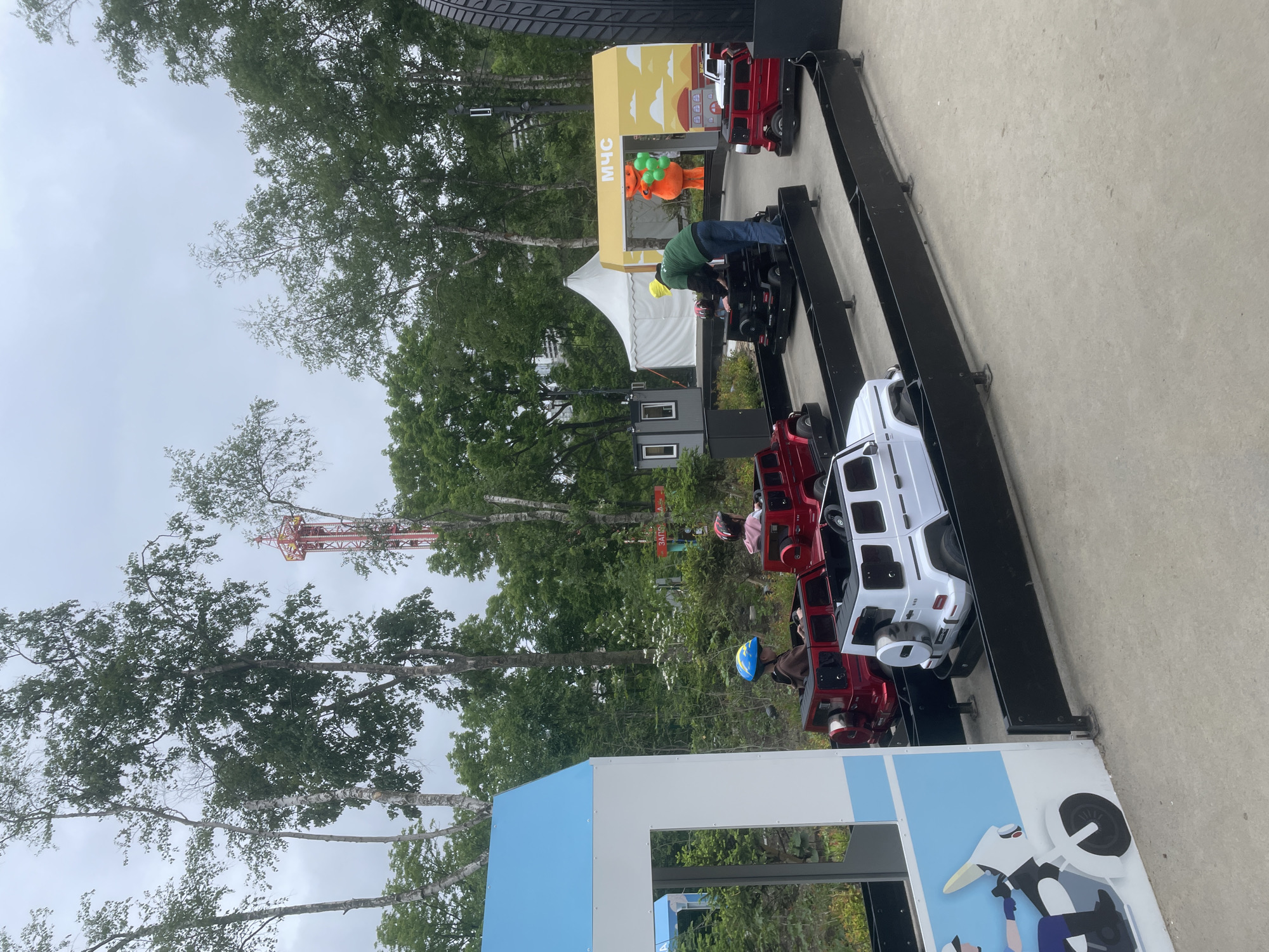 This image captures an outdoor children's driving course within a park-like setting in Vladivostok, Russia. The scene depicts a themed amusement area, likely at the "Sputnik-Vostok" amusement park, given the visible red tower structure in the background.
In the foreground, a light gray concrete surface serves as the base for a miniature vehicle track defined by low black barriers. Several white and red replica Mercedes-Benz G-Wagen toy cars are positioned on this track. On the left, a blue and white structure resembling a house or a police station features a graphic of a police officer on a motorcycle. Two children are seated in red toy cars; one child, wearing a blue and yellow helmet, is visible on the left, while another, wearing a pink helmet, is further back. An adult male, dressed in a green long-sleeved shirt, blue jeans, and a yellow cap, is bent over a black toy car on the track, seemingly assisting a partially obscured child.
In the midground, to the right, a yellow archway structure is visible, emblazoned with the Cyrillic text "МЧС" (MChS), indicating the Ministry of Emergency Situations, along with cartoon depictions of buildings and clouds. An orange mascot figure, holding a bunch of green balloons, stands under this archway. Behind the driving course, a large white tent and several smaller, dark gray modular kiosks or buildings are present. A red sign in the distance displays the Cyrillic word "ЗАПРАВКА" (ZAPRAVKA), meaning "fuel station" or "refueling."
The background consists of dense natural foliage, dominated by numerous tall deciduous trees, including several with pale bark resembling birch trees. The sky above is overcast and uniformly light gray. Prominently rising above the treeline in the distance is a tall, slender red tower structure, characteristic of a drop tower ride, further confirming the amusement park context. A large black tire, possibly a decorative prop, is partially visible on the far right edge of the frame.