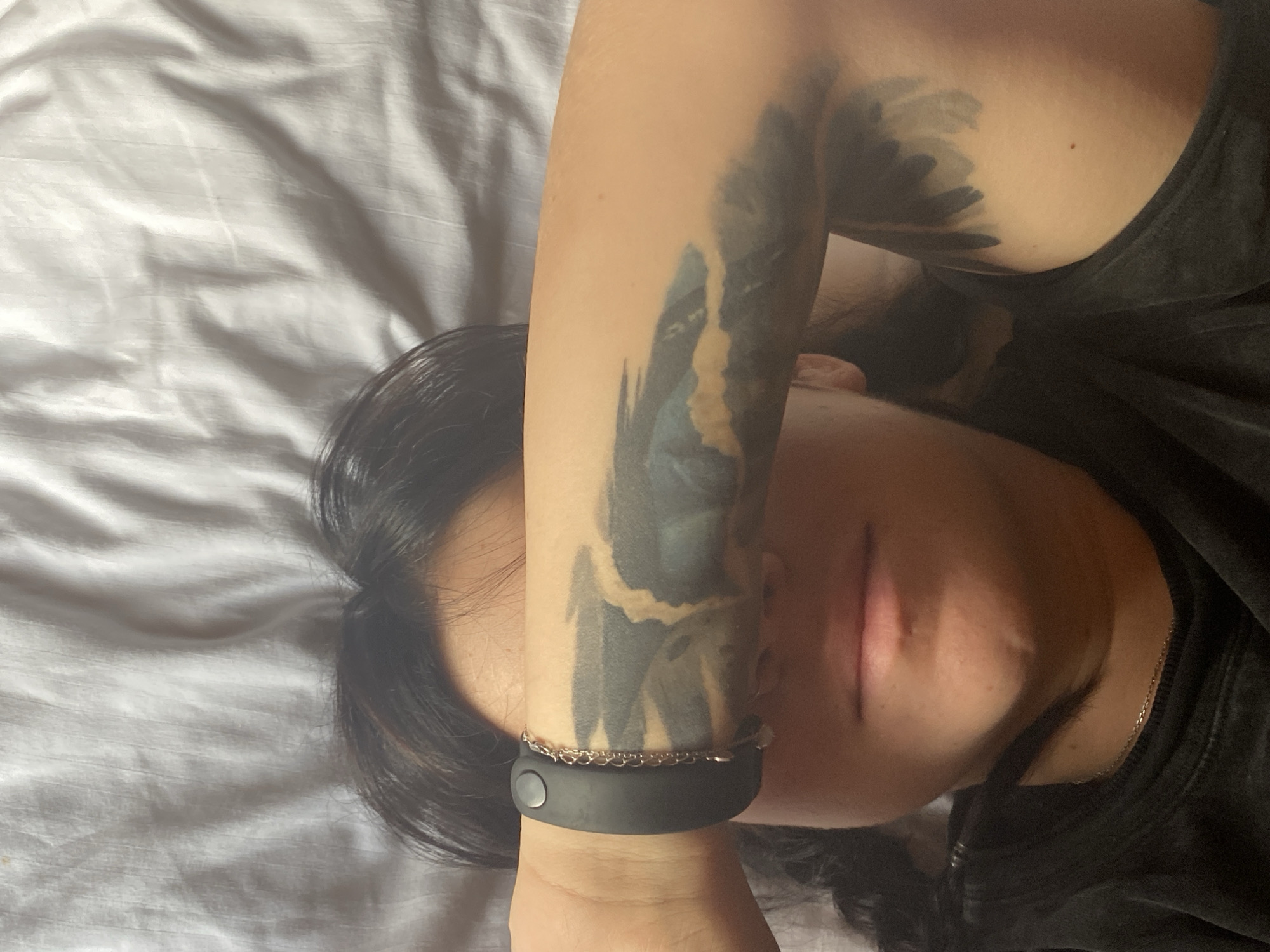 An overhead, close-up shot shows a person lying on a white, wrinkled sheet. The person is wearing a black t-shirt and has dark hair. A large, dark tattoo covers the person's left arm, appearing to depict a landscape with mountains and clouds. A black fitness tracker with a chain bracelet is worn on the left wrist. The person's face is partially visible, with their head tilted and eyes closed. The lighting suggests it is daytime, possibly indoors. There is no discernible information to place this scene in Krasnoflotskoe, Russia.