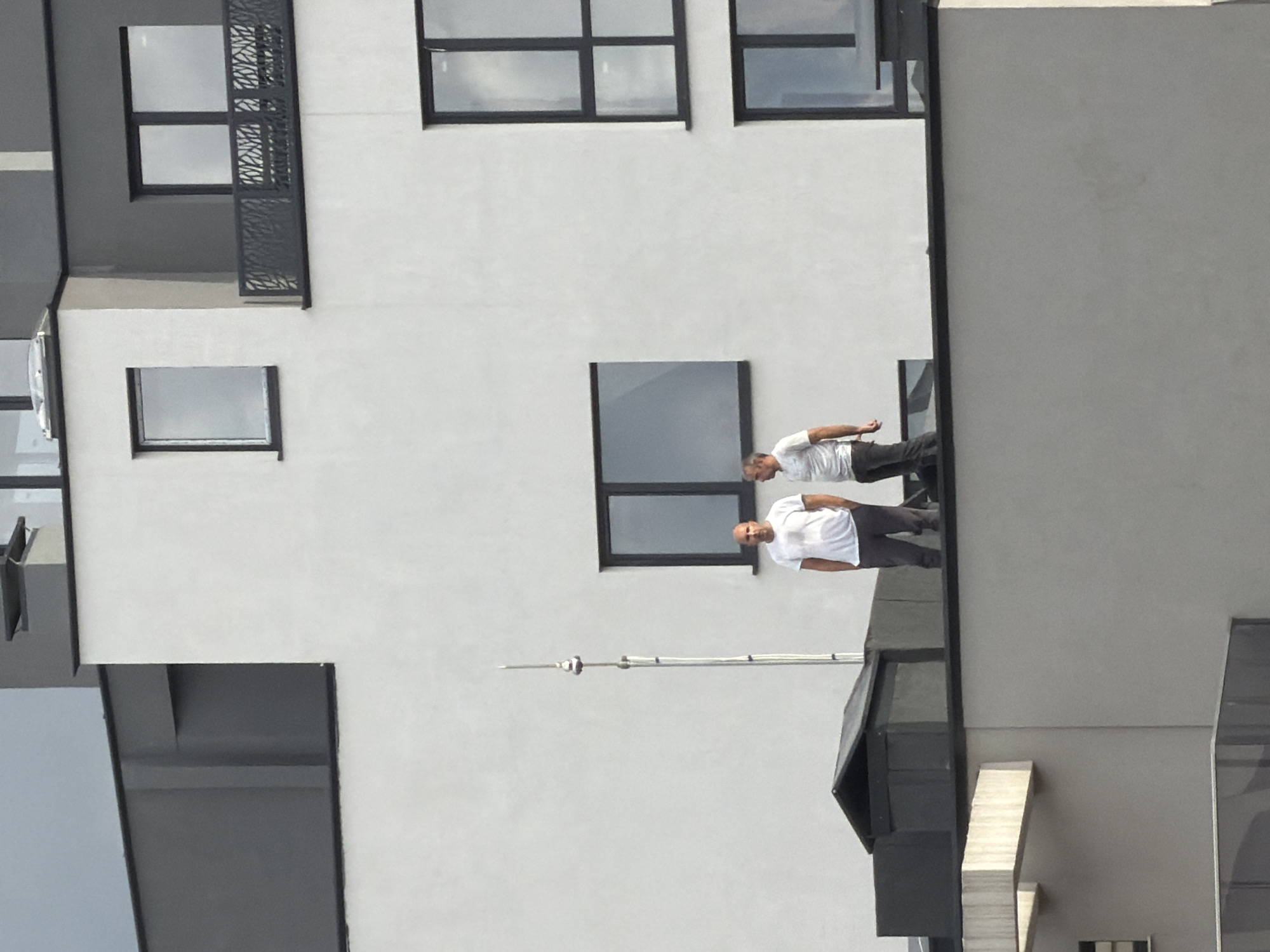 The image depicts the exterior of a modern building, likely located in a city (identified as Sofia, Bulgaria). The building is primarily white with dark window frames. It features several square windows on the facade. There are two men standing on what appears to be the roof of a lower section of the building. Both are wearing white shirts and dark pants. They are positioned close to one of the windows. A thin metal pole, possibly a lightning rod, extends upwards from the lower section of the building. The overall scene is well-lit, suggesting a daytime setting. The photograph is taken from a lower vantage point, looking upwards towards the building and the men on the roof.