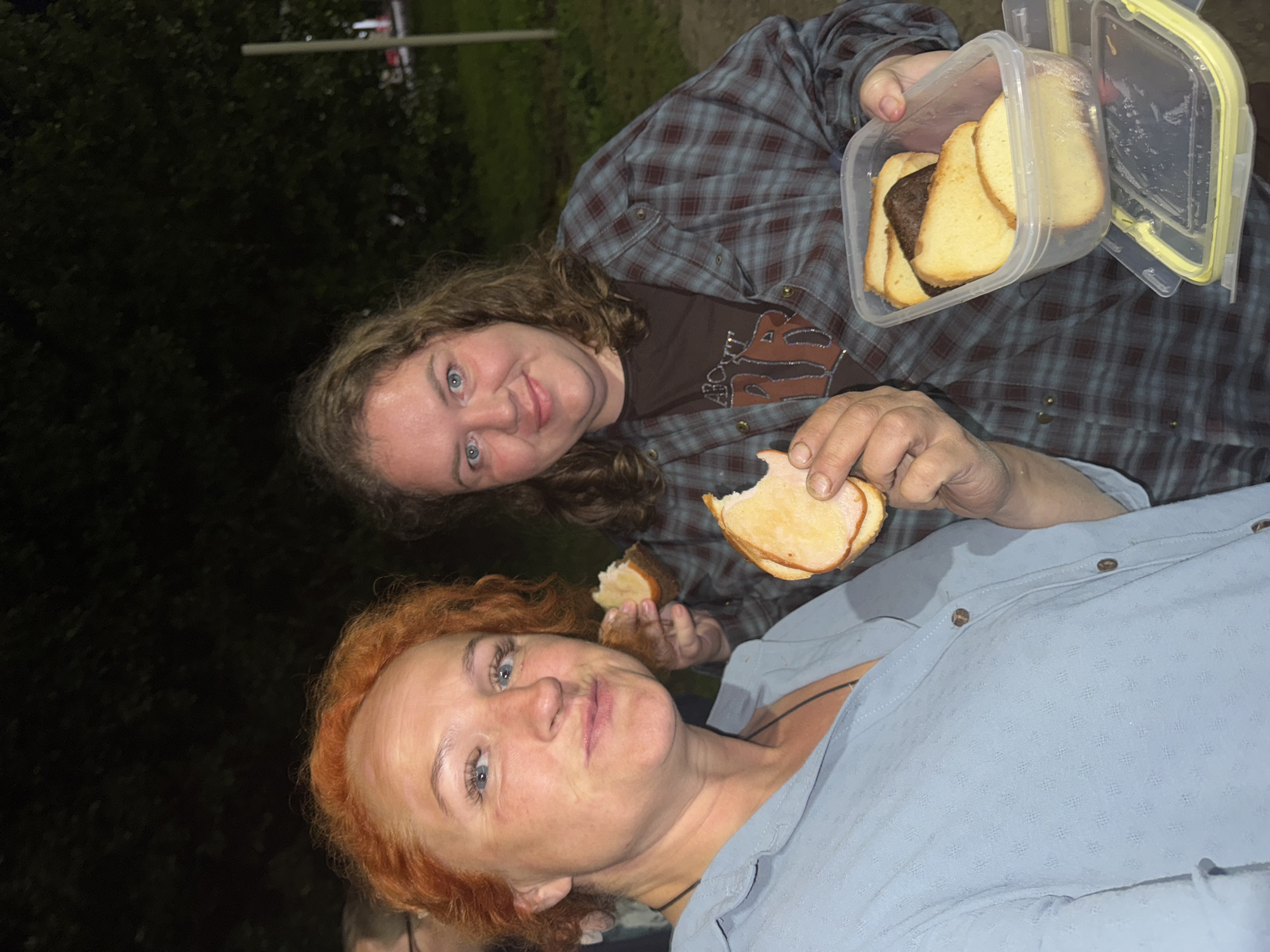 Two women are standing outside at night, possibly in the city of Anatas, Russia. The woman on the left has bright orange hair and is wearing a light blue, collared shirt. She is holding a sandwich with ham and bread up to her face, and appears to be taking a bite. The woman on the right has curly brown hair and is wearing a dark plaid shirt over a brown t-shirt with a graphic design. She is holding a clear plastic container filled with slices of bread and one slice appears to have a dark topping. The background is dark and out of focus, with some trees visible. The lighting is dim, suggesting it is nighttime.