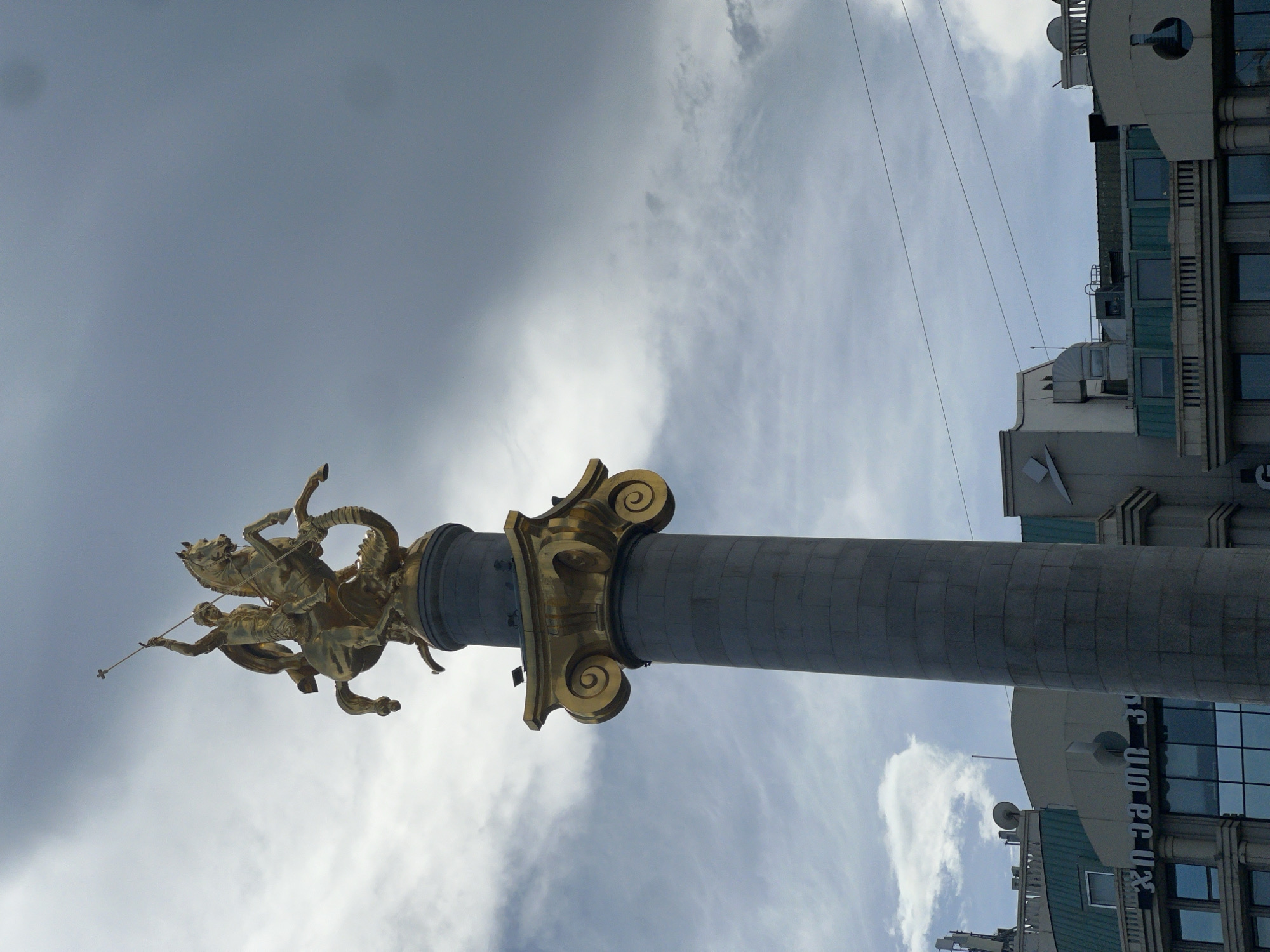The image depicts the St. George Statue in Tbilisi, Georgia, from a low-angle perspective, pointing upward. The statue, featuring St. George on horseback spearing a dragon, is gilded and sits atop a fluted gray stone column with a gold-colored Corinthian capital. The sky is overcast with a mix of dark and light clouds. To the right of the column are parts of nearby buildings, including glass windows, architectural details, and satellite dishes. Power lines are also visible in the upper right corner of the frame.