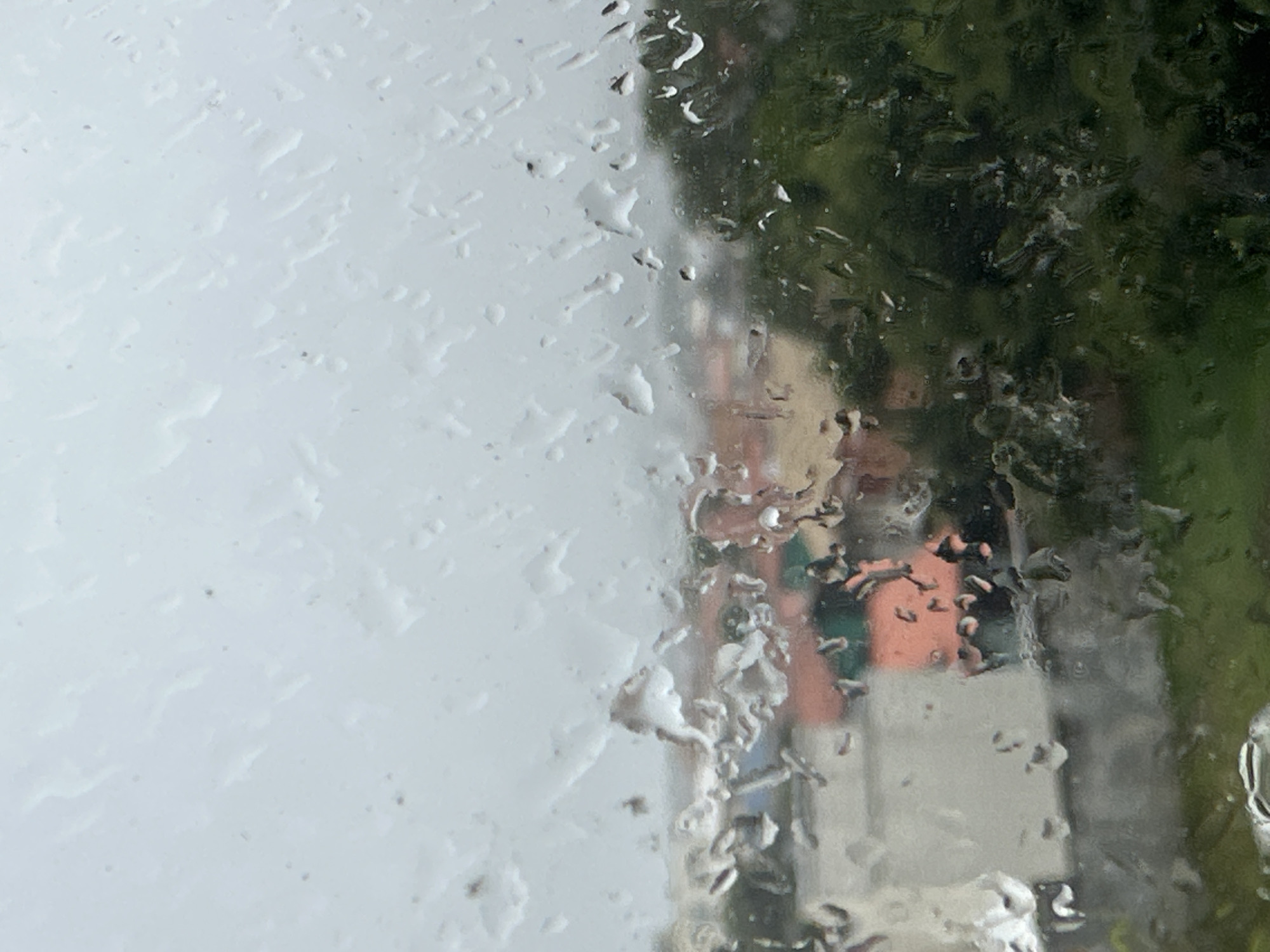 The image shows a view through a rain-streaked window, looking out at a blurred urban landscape. The foreground is dominated by numerous raindrops of various sizes clinging to the glass, distorting the view behind. Beyond the window, indistinct buildings with reddish-orange and white facades are visible, nestled amongst lush green trees. The sky is overcast and appears bright white, suggesting a cloudy or rainy day. This scene is characteristic of the typical weather experienced in Florianópolis, Brazil, a coastal city known for its frequent rainfall, especially during certain seasons. The overall impression is one of a somber, subdued atmosphere, common during periods of inclement weather in this vibrant Brazilian city.