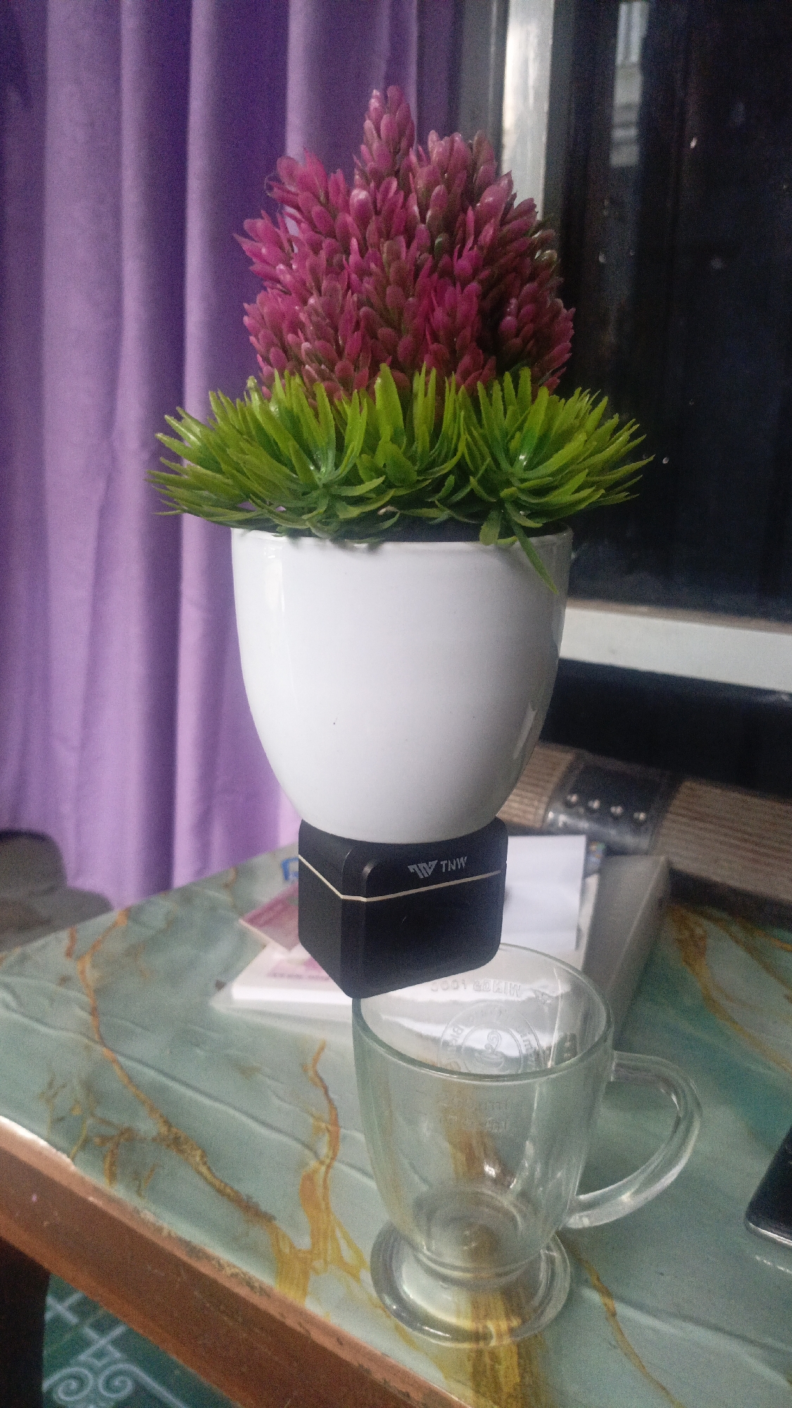 A potted artificial plant with pink and green foliage sits atop a black electronic device labeled "TNW" which is precariously balanced on a stack of papers. Beneath this arrangement, a clear glass mug with a handle rests on a table with a green and gold marbled surface. In the background, a purple curtain hangs. The scene appears to be indoors, possibly on a desk or table. The image was taken in Salatiga, Indonesia.