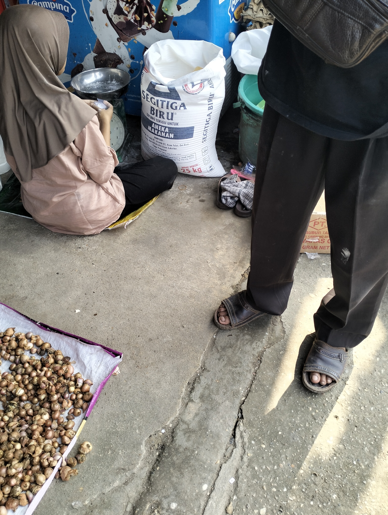 In Serdang Jaya, Indonesia, a market scene unfolds with two individuals and various goods. A woman, facing away from the camera, is seated on a yellow mat, wearing a light brown hijab and a matching colored blouse with black pants. She is engaged in weighing something on a green scale, with a silver bowl placed on top of it, and appears to be holding a small white item. Beside her, to her right, is a large white bag of "SEGITIGA BIRU TEPUNG TERIGU UNTUK ANEKA MAKANAN" (Three Blue Triangle Wheat Flour for Various Foods), which states it contains 25 kg. Further to the right, a person stands, wearing dark pants and dark leather sandals. Their left leg is partially visible, and they appear to be carrying a dark leather bag or satchel over their shoulder. In the foreground, on the left, a collection of small, dried bulbs, possibly garlic or shallots, are spread out on a white surface with purple trim. The ground is concrete, showing cracks and imperfections, with a shadow cast by the standing person falling across it. A green plastic bucket is visible behind the flour bag. The overall environment suggests an outdoor market setting with a focus on food-related items.