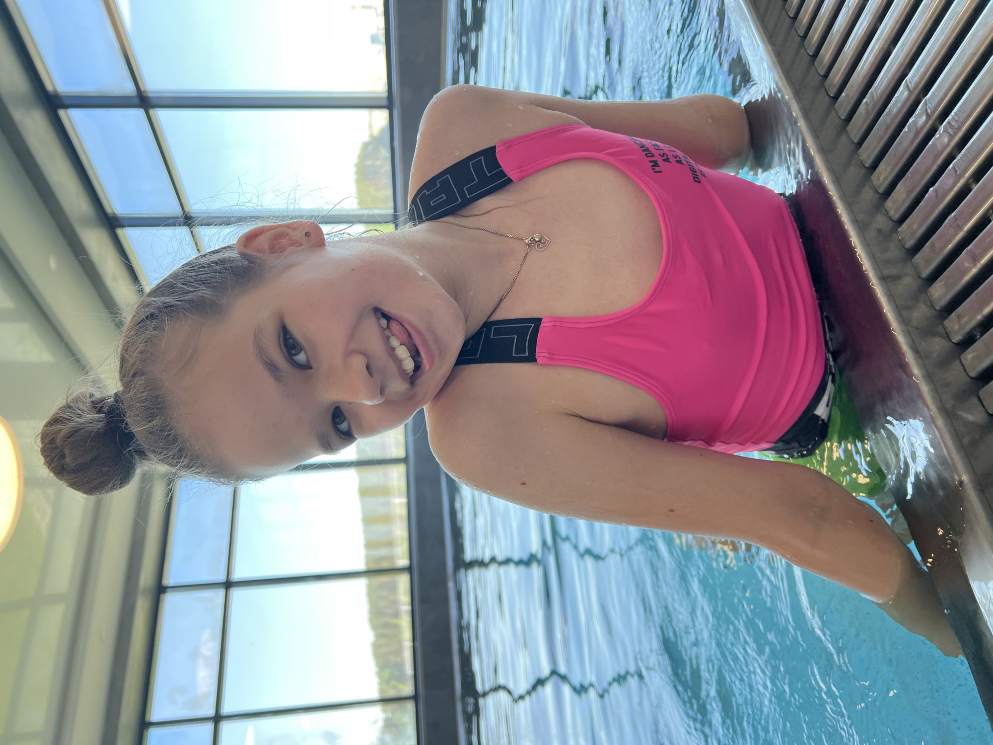 A young girl is partially submerged in a swimming pool, wearing a pink swimsuit. She is leaning against the side of the pool and smiling at the camera. The pool is indoors, as evidenced by the window panels in the background. The water appears clear and blue. The location is the city of Chaikovsky, Russia.