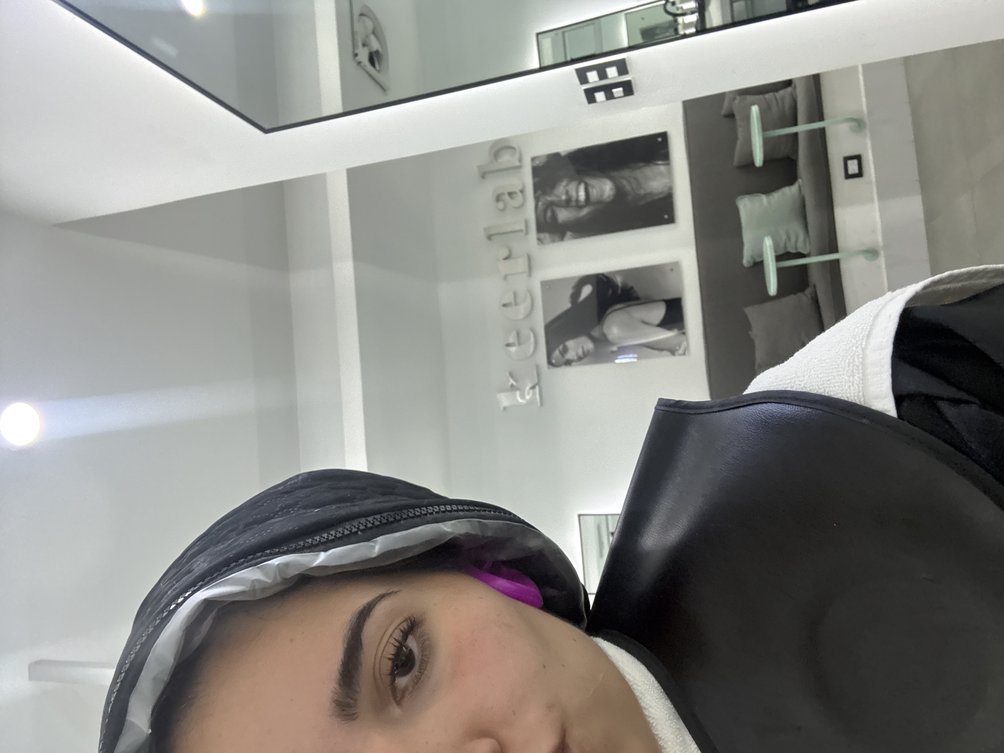 The photograph appears to be a selfie taken indoors. A person with dark hair and visible eyelashes is wearing a black hood with a light gray lining and a purple ear covering. The individual is partially obscured by what seems to be a black protective garment, possibly leather, over a white towel.
In the background, the "kerlab" sign is displayed vertically with two black and white photos placed above a gray seating area with light green support structures and pillows. The environment looks clean and modern, possibly a salon or spa.
The photograph does not provide information to confirm the location in the city of San Bernardino Tlaxcalancingo, Mexico.