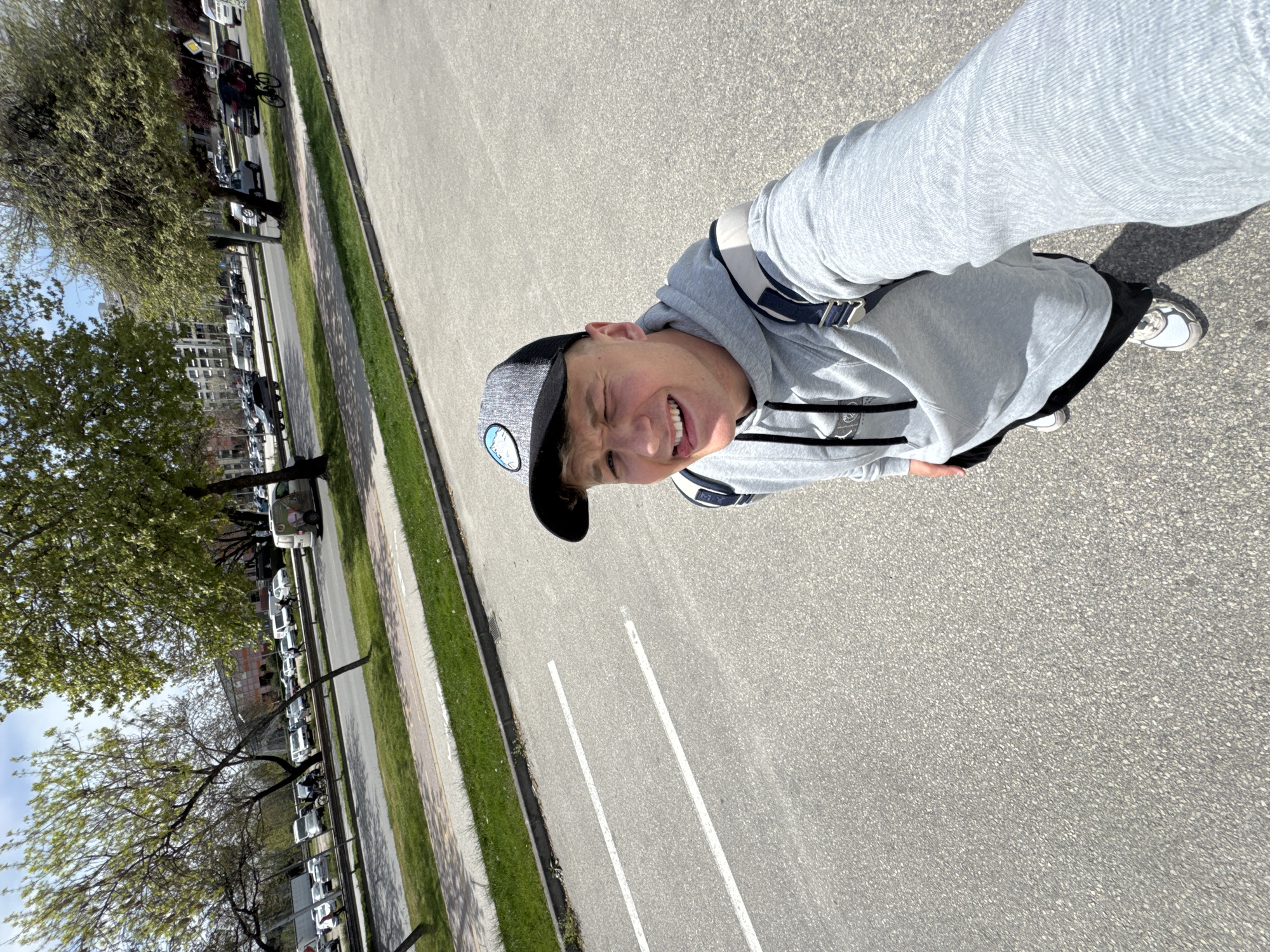 The image captures a playful moment featuring a young man outdoors on a sunny day in Plovdiv, Bulgaria. He is the central subject, looking directly at the camera with a wide, open-mouthed smile, his tongue slightly out, conveying a cheerful and energetic mood. He wears a grey hooded sweatshirt with black and white striped accents on the sleeves, a grey and black baseball cap with an unclear circular logo on the front, and white sneakers with dark details. A dark strap, likely from a bag, is visible across his chest.
The setting appears to be an urban or suburban street. To the left, a line of trees with fresh green leaves suggests spring or early summer. Below the trees are a sidewalk and a strip of grass. Beyond these, a row of parked vehicles, including what looks like a white van with graphics, lines a road. Buildings are visible in the distance through the trees. The right side of the frame features a wide expanse of asphalt with distinct white line markings, possibly indicating parking spots or traffic lanes. The bright sunlight casts strong shadows, confirming clear weather conditions during the daytime. The photograph is taken from a slightly elevated, tilted perspective. No visible text is clearly legible on any objects within the image.