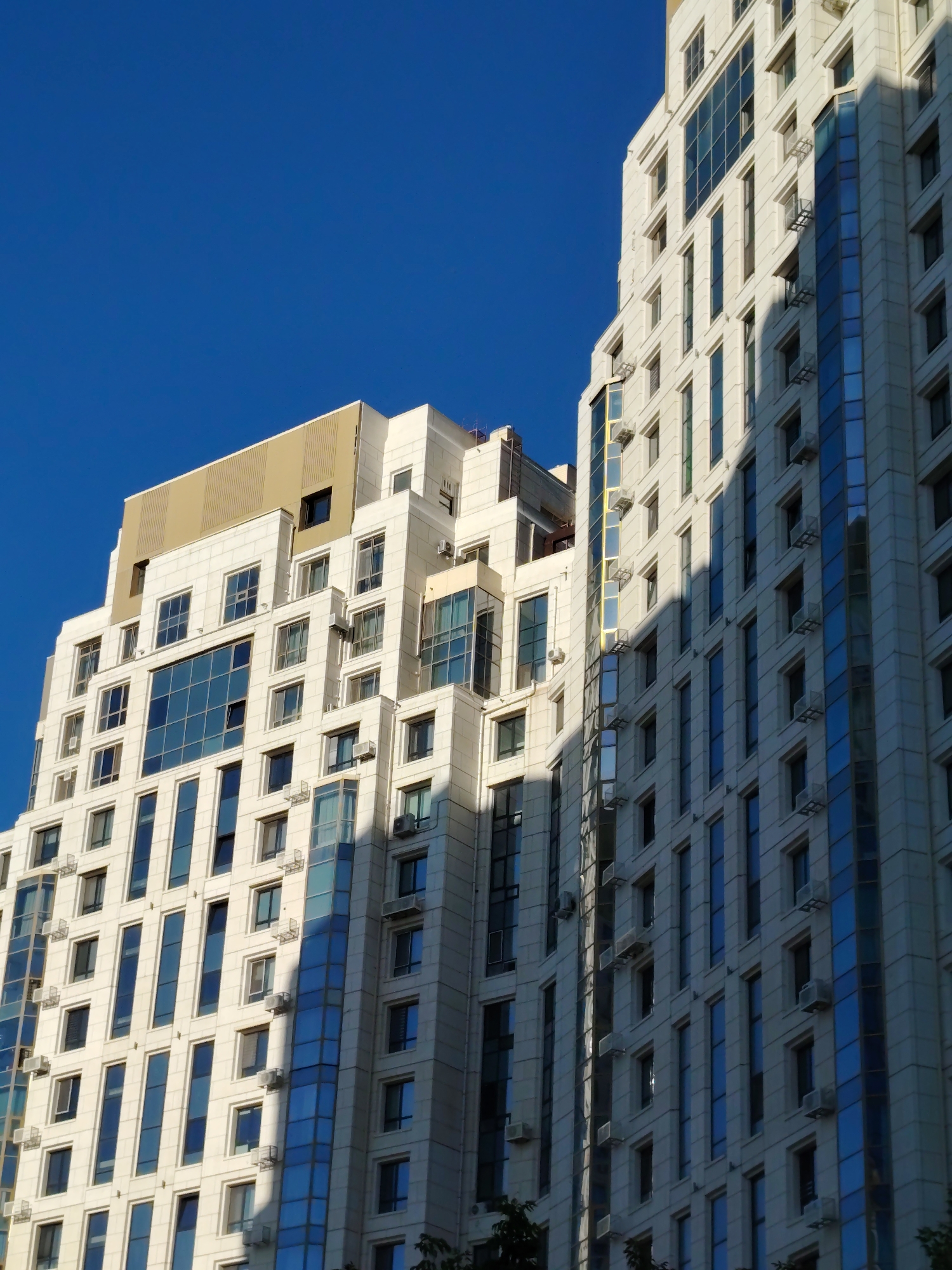 The image shows a modern residential building with a light-colored facade, large windows, and balconies. The building appears to be a multi-story structure with a stepped design. Some windows are reflective, showing the blue sky. The building is casting a shadow due to the sunlight. The sky is clear and blue. The architectural style is contemporary, typical of urban development. This building is located in Astana, Kazakhstan.