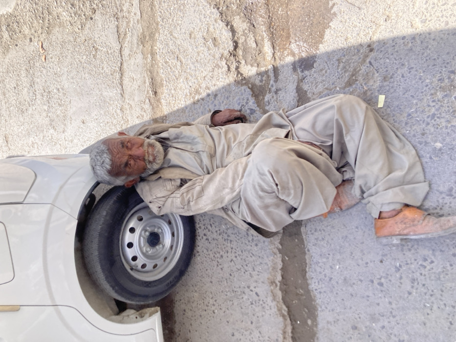 An elderly South Asian male is seated on a cracked, uneven concrete or asphalt surface. He wears a worn, light-colored shalwar kameez and light brown shoes. The man presents with grey hair and a full grey beard, deep-set eyes, and a wrinkled facial structure, looking directly towards the viewer. His posture involves leaning against the rear passenger-side wheel and lower body panel of a white vehicle, likely a van or mini-truck, which is partially visible on the left side of the frame. Strong daylight casts defined shadows across the scene. A small, light-colored rectangular object lies on the pavement near his left leg. The specific location within Quetta City Tehsil, Pakistan, is not discernible from the visual evidence.