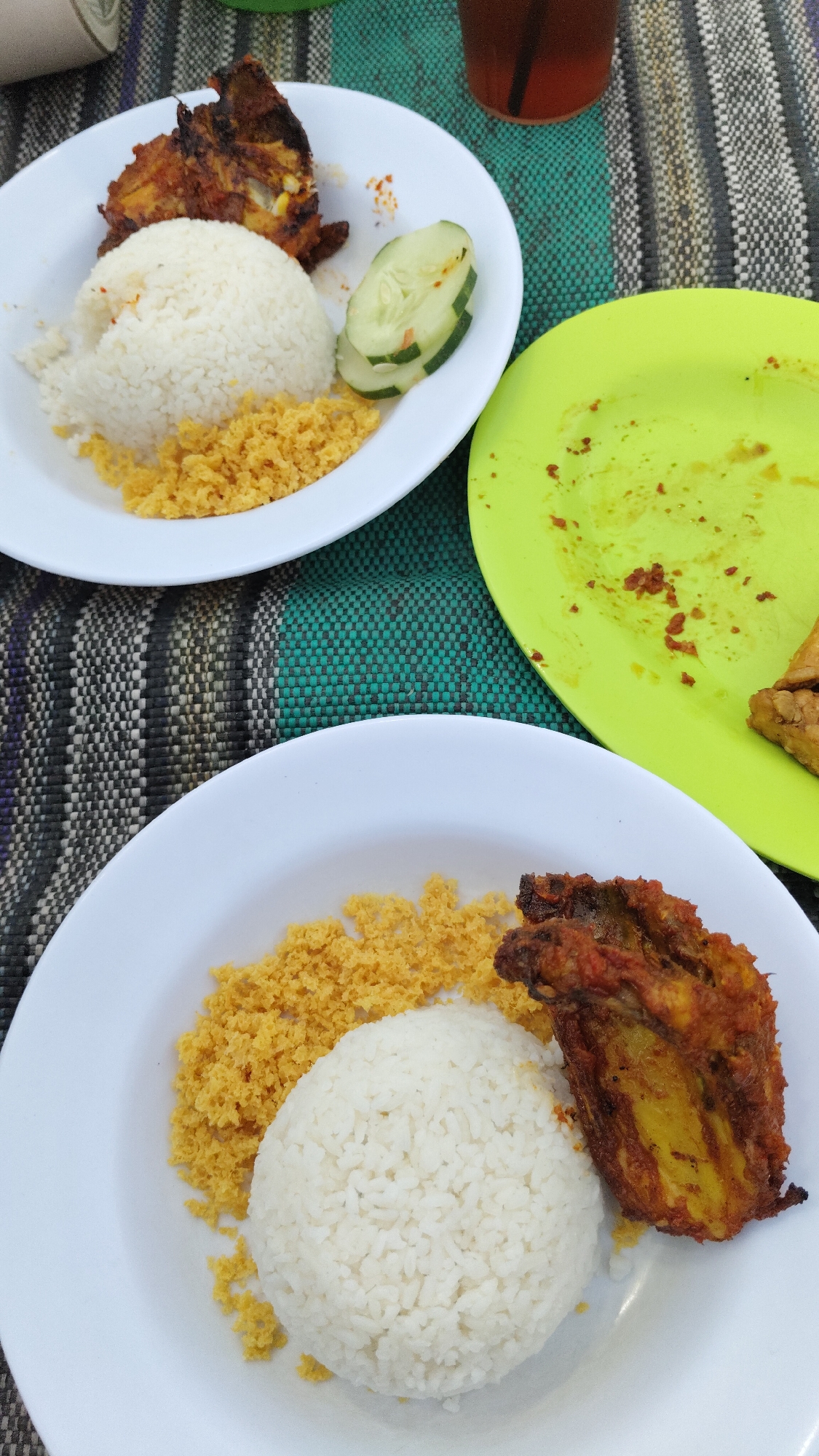 An overhead, close-up shot shows two plates of food on a woven placemat. The plate in the foreground contains a mound of white rice, a portion of golden-brown fried chicken, and a scattering of what appears to be fried crumbs or spiced rice. The plate in the background, positioned above and to the left of the foreground plate, holds a similar serving of rice and fried chicken, accompanied by a few slices of cucumber. A green plate with remnants of food is visible to the right of the background plate. A dark beverage in a clear glass with a straw is situated at the top right of the image, partially obscured by the placemat. The overall impression is of a casual meal, possibly at an outdoor eatery in Madiun, Indonesia, given the visual cues of local cuisine and the rustic placemat. No individuals are visible in the frame.