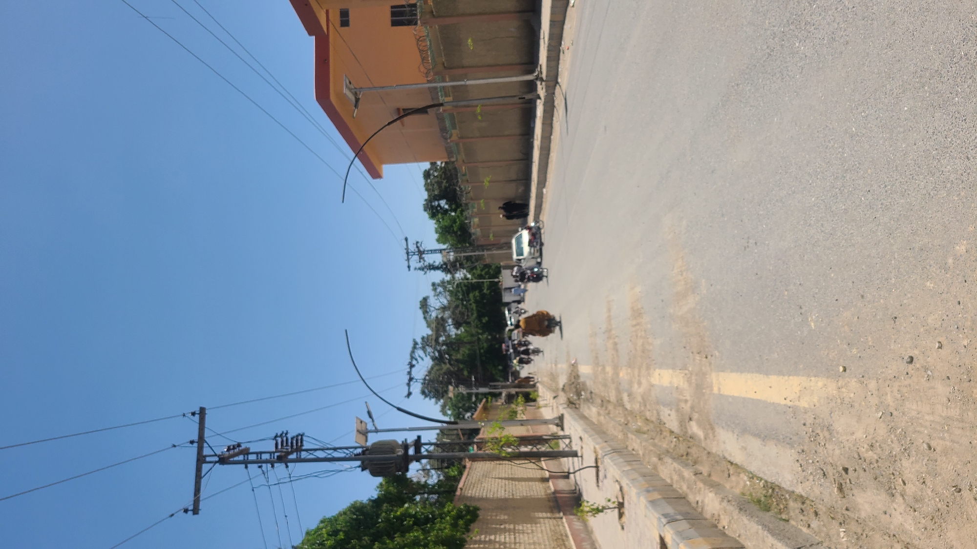 The image shows a street in Quetta City Tehsil, Pakistan. The sky is blue. Buildings are along one side of the street with electrical poles and wires. There are people on motorcycles riding on the street. The road surface is paved. There is some vegetation along the side of the road.