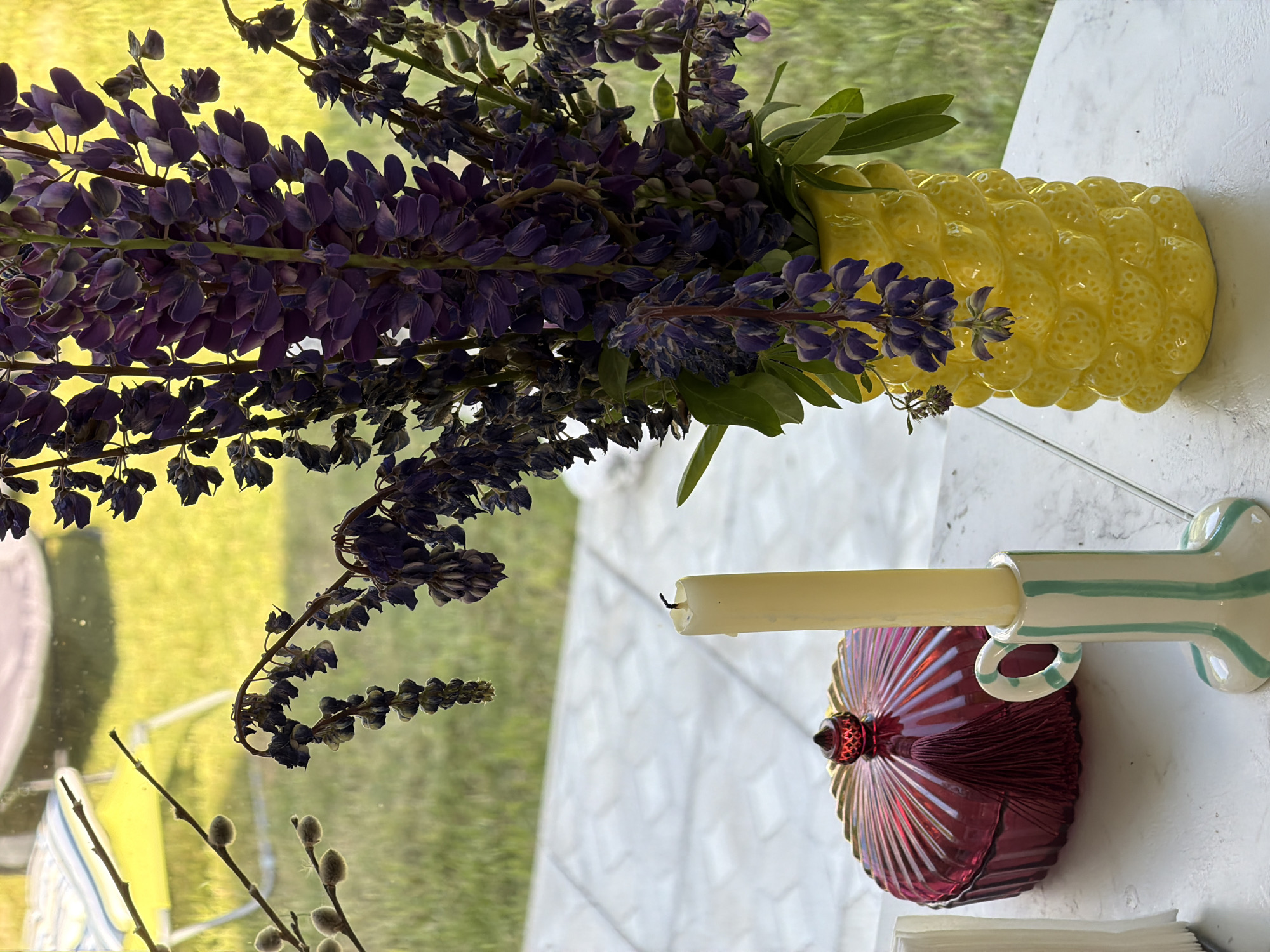 The image depicts a still life arrangement on a white table with a tiled outdoor background. In the foreground, a tall, unlit cream-colored candle stands in a small, striped ceramic candle holder with a light green and white stripe pattern and a loop handle. To the left of the candle is a decorative, deep red glass dish with a fluted lid and a small, faceted red finial on top. A dark red tassel hangs from the lid.
Behind these objects, a bouquet of dark purple lupine flowers is arranged in a bright yellow, textured ceramic vase that resembles a pineapple. The flowers are mostly in bloom, with some withered blossoms visible. Green leaves are interspersed with the flowers.
Through a window or glass partition in the background, a grassy area with blurred green and yellow tones is visible. A yellow chair with a striped cushion is also discernibly out of focus. The tiled ground in the background suggests an outdoor patio or walkway. There are no people or animals present in the image. The setting appears to be a domestic outdoor space, potentially a patio or balcony, in or near Алешкино Дп, Russia.