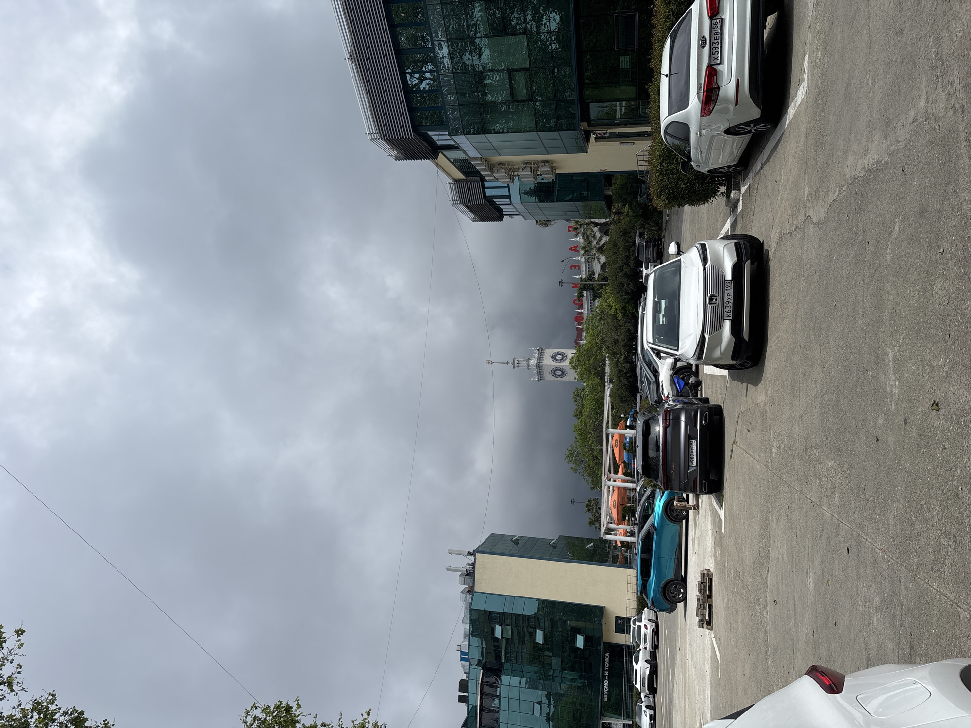 The image depicts a street scene in Sochi, Russia, identifiable by the architectural style and the presence of the Sochi train station in the background. Overcast weather conditions create a subdued atmosphere. The foreground showcases a parking area with various parked cars, including white, black, and turquoise vehicles. Buildings with modern glass facades line the street, and street furniture like lamp posts and umbrellas are visible. Power lines stretch across the sky. The street is situated in close proximity to the city's railway station, which contributes to the overall urban feel of the setting.