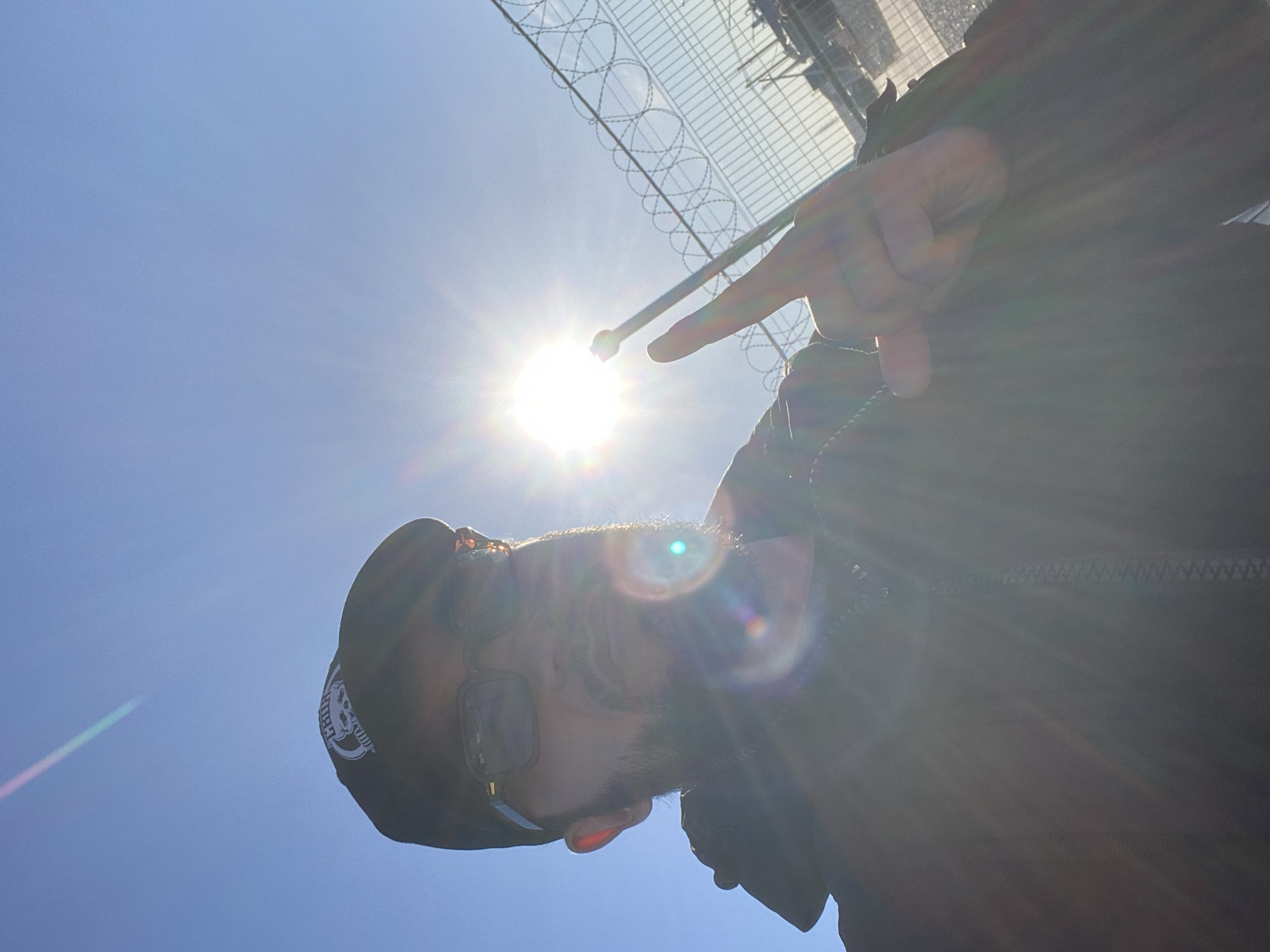 A selfie is taken outdoors on a sunny day. The sun is bright and positioned in the upper right quadrant of the frame, causing lens flare and overexposure. A person, partially obscured by the sun's glare, is visible in the foreground. They are wearing a black baseball cap with a white circular logo featuring a skull. They also wear glasses and have a beard. The person's right hand is raised, and they are making a peace sign with their index and middle fingers. In the background, a metal fence with barbed wire runs horizontally across the frame, separating the viewer from a blurry industrial area with some structures and possibly vehicles. The sky is clear and blue. The location is identified as Koskolovo, Russia.