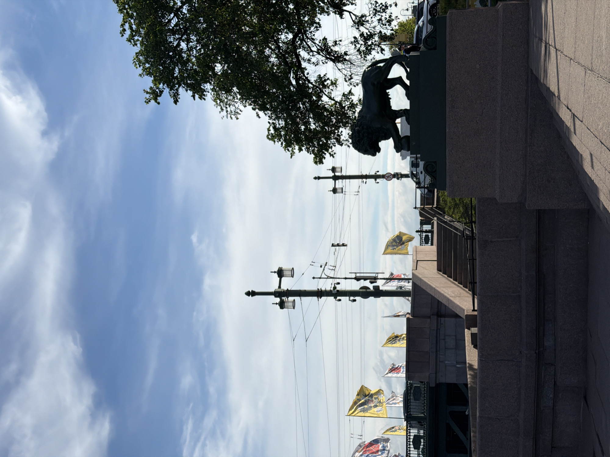 The image shows a bronze lion statue on a granite pedestal, located on a bridge in Saint Petersburg, Russia. The statue depicts a lion standing on a sphere. In the background, there are several lampposts, flags, and overhead wires. A large tree branch hangs over the statue. A few cars and people are visible in the background. The sky is partly cloudy. This is the Lion Bridge (Lviv Most) over the Griboyedov Canal, famous for its lion sculptures.