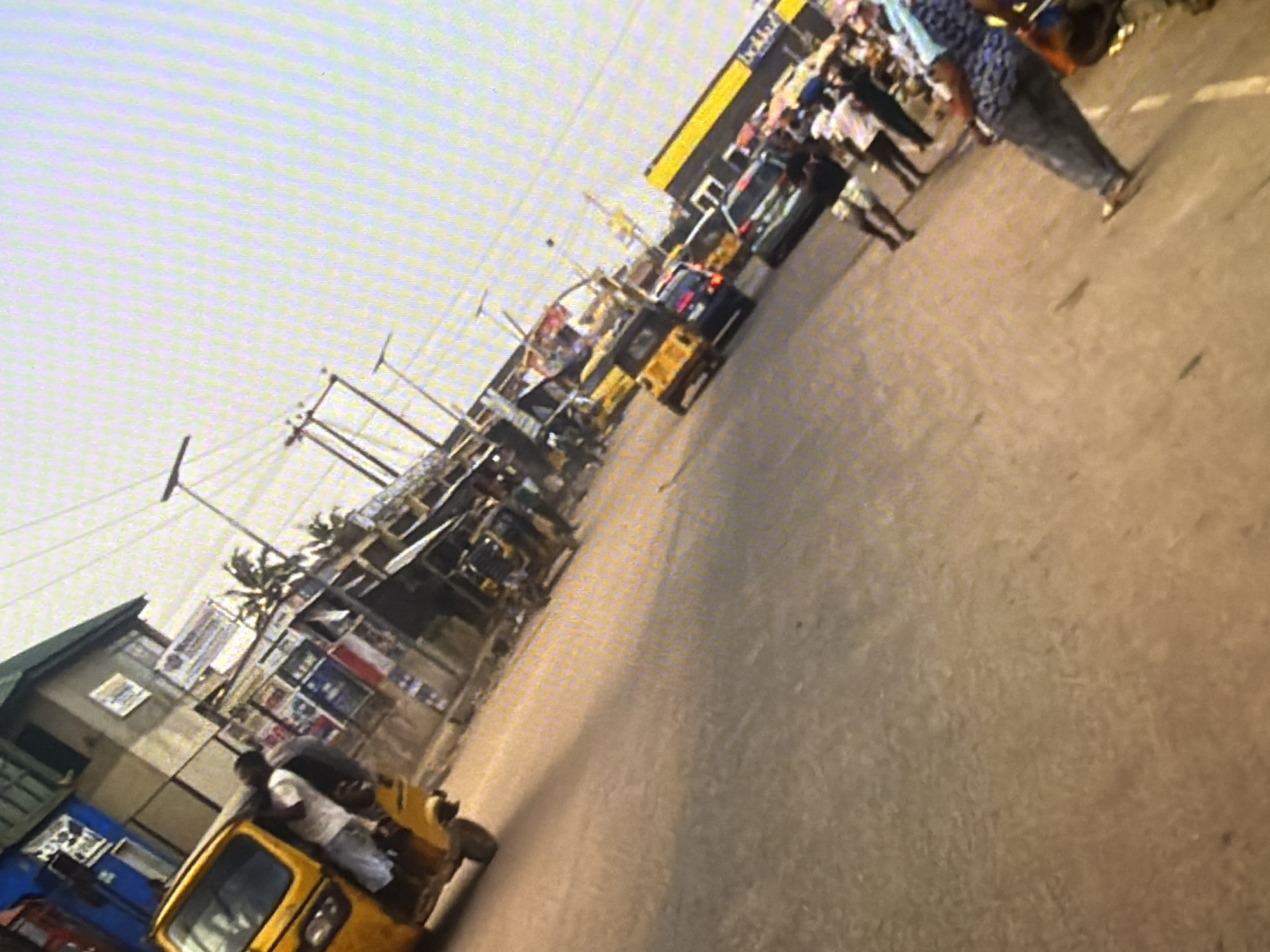 The image depicts a busy street scene, likely in Bukuru, Nigeria. The perspective is from a low angle, possibly from a moving vehicle, as indicated by the motion blur. In the foreground, a yellow motorized tricycle, commonly known as a 'keke napep,' is visible, with a person standing beside it. The street is paved with asphalt and shows signs of wear and tear.
Further down the street, a line of similar tricycles and a few cars are present, suggesting a traffic bottleneck or a busy transport hub. The background features several low-rise buildings with signage, some of which are faded or obscured. The architecture appears typical of a Nigerian urban or semi-urban environment.
Several individuals are seen on the street, some appearing to be passengers or vendors, while others are simply pedestrians. Their clothing is casual, fitting for a warm climate. The overall atmosphere is one of a bustling commercial area, characterized by the presence of various vehicles and people engaged in daily activities. The sky is overcast, and the lighting suggests it is either early morning or late afternoon. The image captures a snapshot of everyday life in a Nigerian city, with a focus on transportation and commerce.