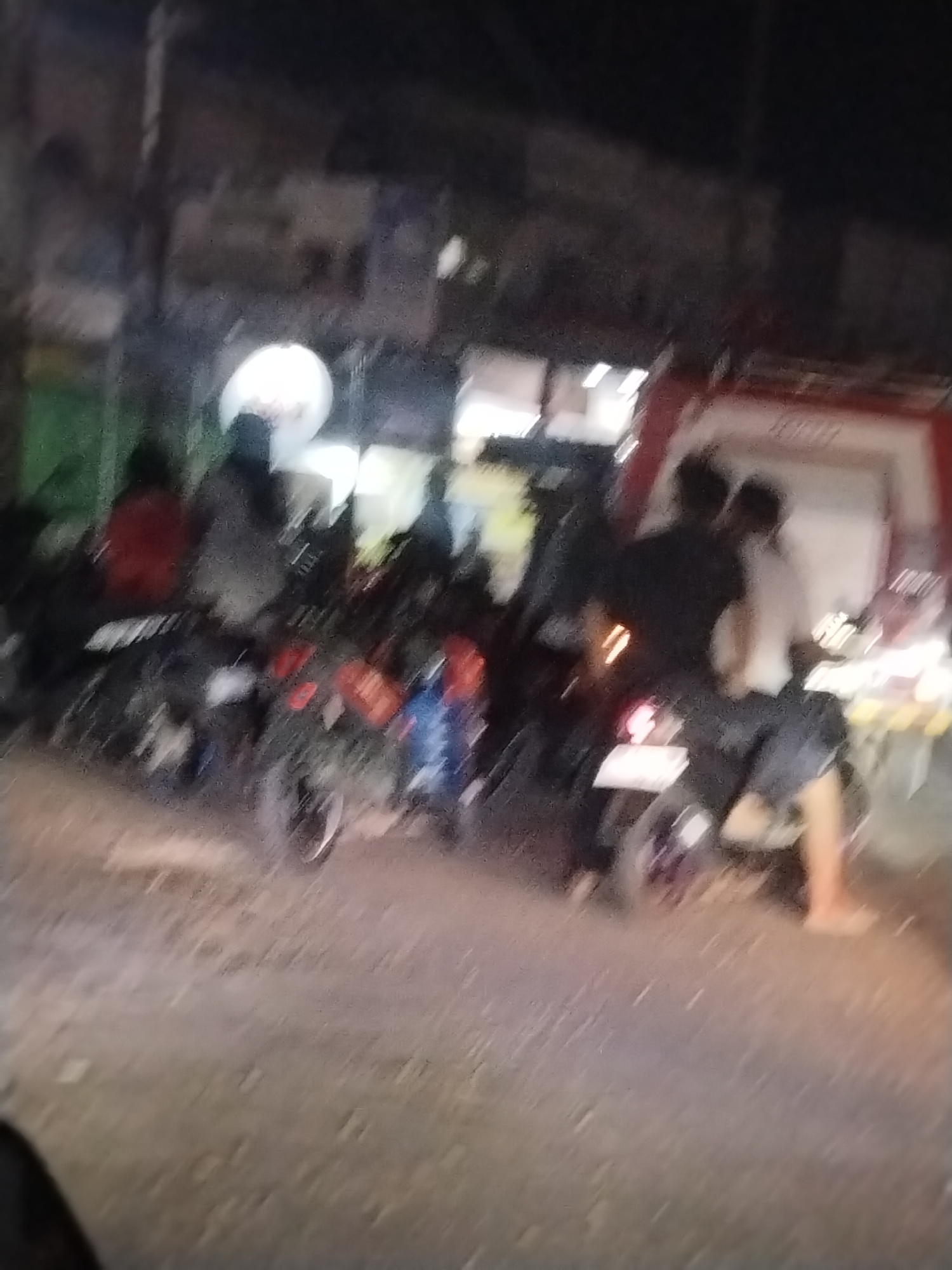 The image depicts a blurry, night-time scene in an urban setting. Several motorcycles are parked or stopped on a paved road, with riders or passengers seated on them. In the background, there are buildings with some illuminated windows and a red structure, possibly a vehicle or part of a building. The overall impression is of a street or parking area at night. The location is identified as Unknown, Indonesia.