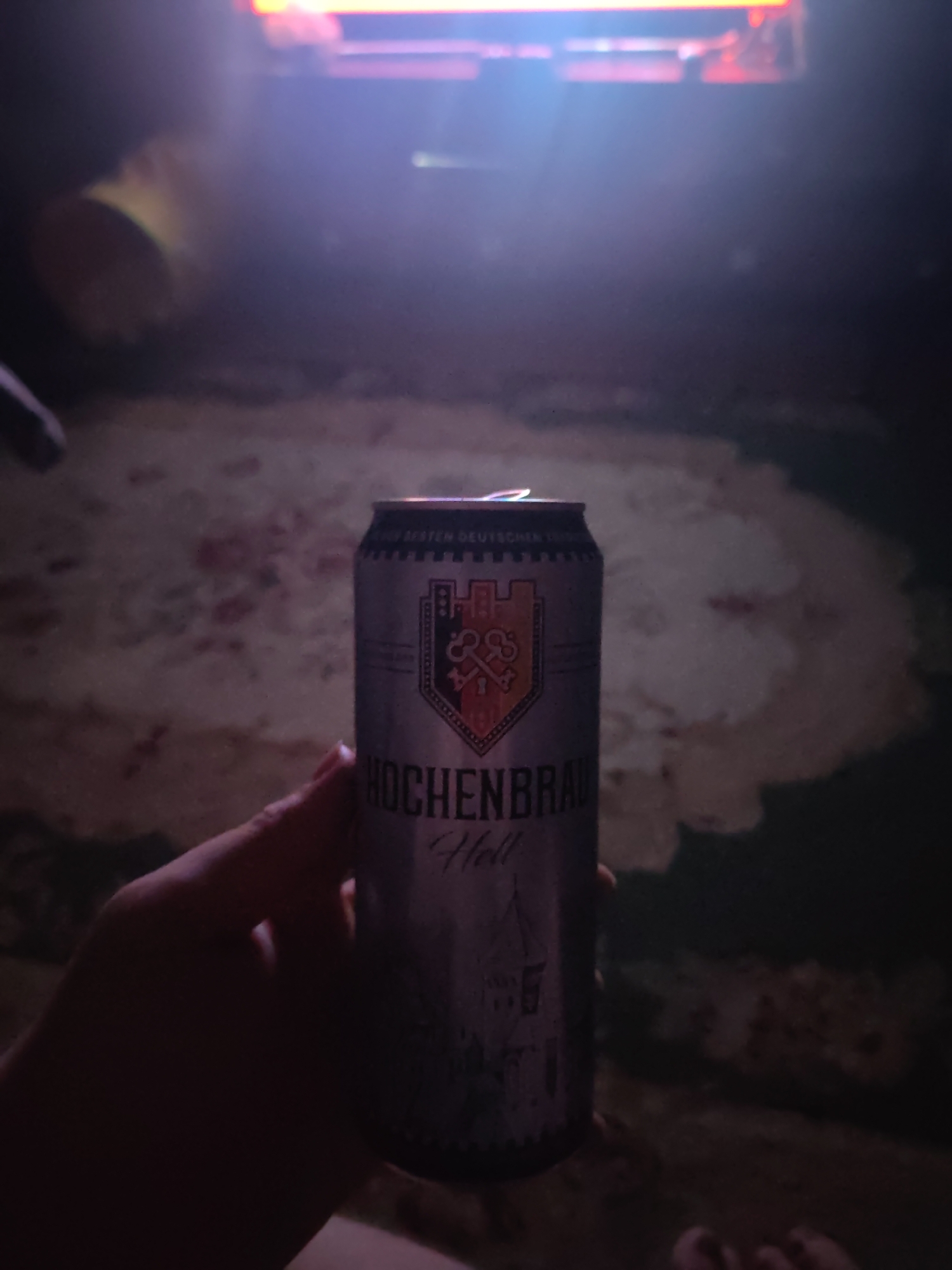 In a dimly lit indoor setting, a person's hand holds a silver can of "HOCHENBRAU Hell" beer. The can features a coat of arms with a castle and keys, along with text that is partially obscured. The background is indistinct, showing a pattern on what appears to be a rug or carpet and a blurry light source at the top, possibly a screen or lamp. There are no discernible elements in the image that would suggest a specific location in the city of Vladimir, Russia, or any urban context. The scene is primarily focused on the can of beer and the hand holding it.