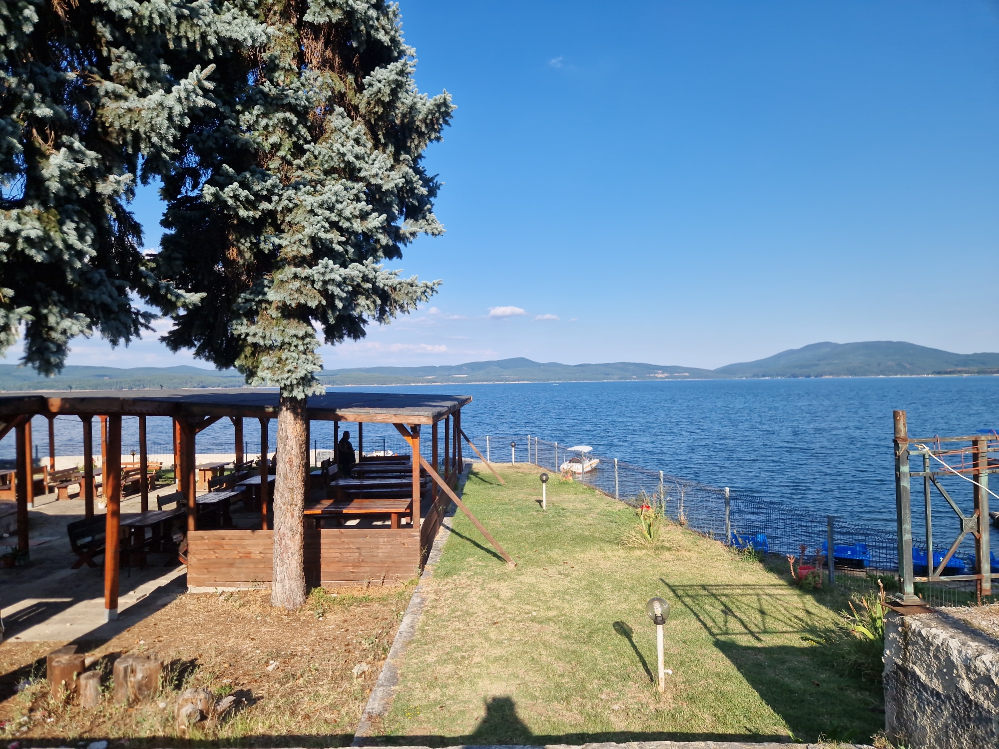 The image depicts a waterfront outdoor restaurant or seating area by a large body of water, likely the Iskar Dam near Dolni Pasarel, Bulgaria. In the foreground, a large evergreen tree with blue-green needles dominates the left side of the frame, casting shadows on the ground. To the right of the tree, a wooden structure with a covered roof supported by thick wooden posts provides shade over several picnic tables and benches. One person is visible in silhouette, seated at a table within this structure. Beyond the wooden shelter, a well-maintained lawn stretches to the edge of the water. A wire fence runs along the perimeter of the lawn, separating it from the water. Several small, blue paddle boats are visible moored in the shallow water near the shore. In the distance, a small white boat is anchored on the water, and a hazy mountain range forms the horizon under a clear blue sky with a few wispy clouds. The scene suggests a peaceful, recreational environment.
