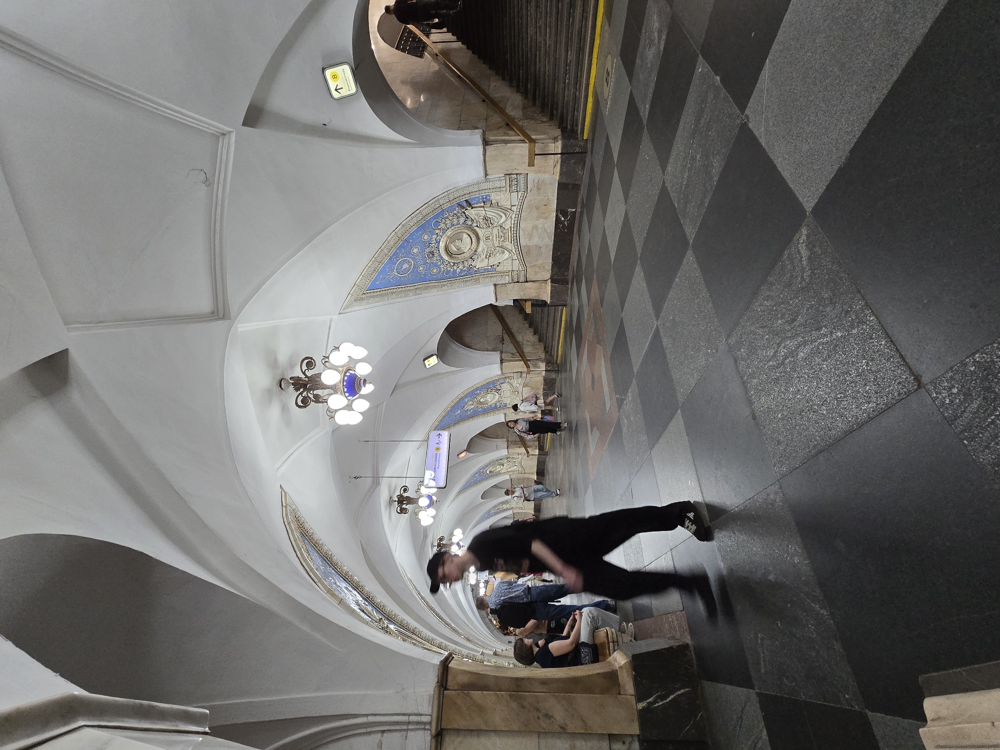 A rotated, eye-level, wide-angle shot depicts a hallway with people in an unknown location in an unknown city. The floor is tiled in a black and gray checkerboard pattern. The walls and ceiling are white, with arched ceiling elements adorned with decorative blue and gold mosaics. Several ornate chandeliers hang from the ceiling. People are scattered throughout the hallway. A man in black clothing is running in the foreground. Several people are sitting on the ledge on the lower left of the frame. The background reveals a staircase leading upwards. A yellow sign with a black arrow is visible on the left side of the frame.