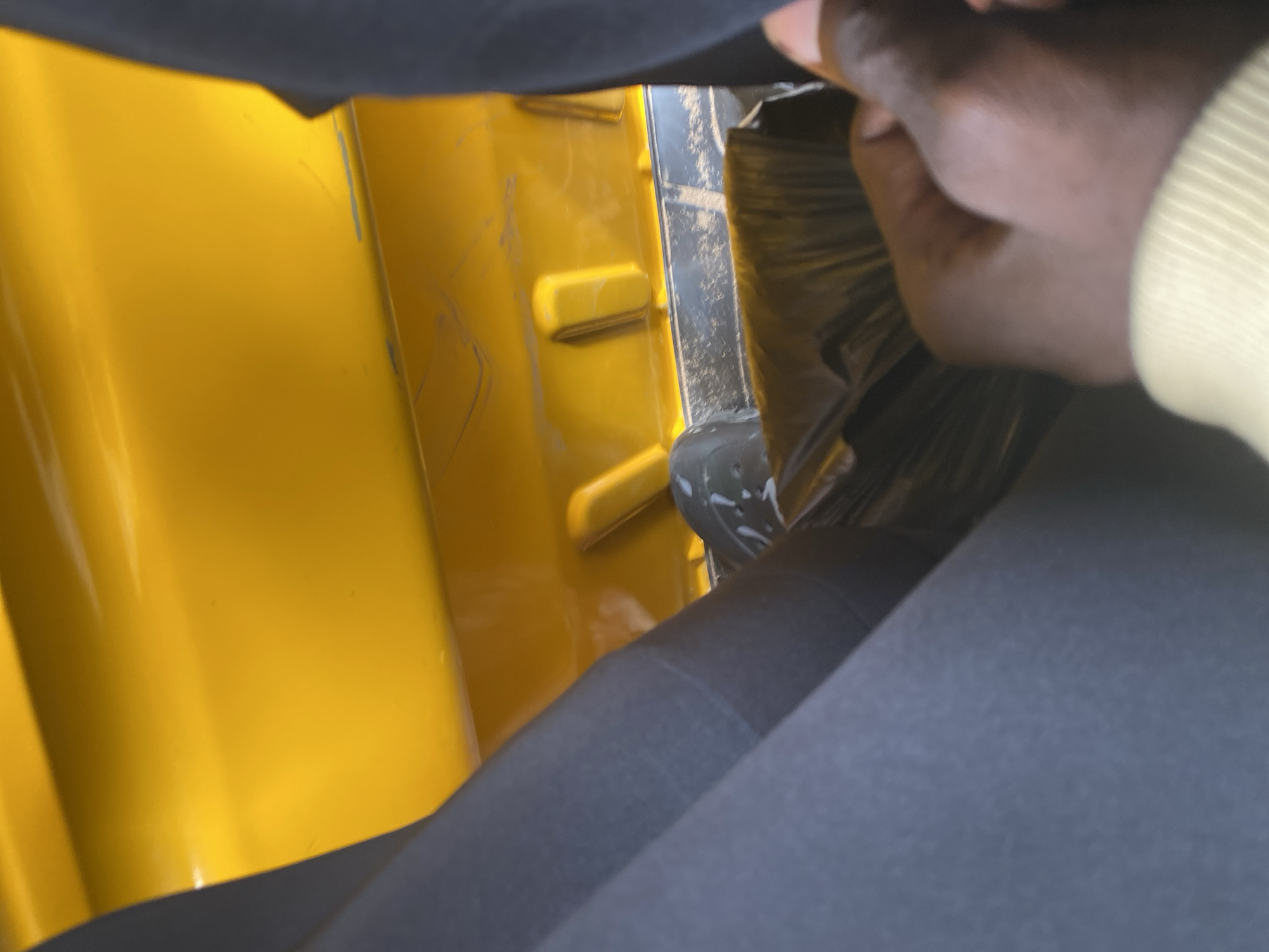 The image shows a person's hand reaching into a black plastic bag, which appears to be inside a yellow container. The person's hand is dark-skinned, and they are wearing a light-colored sleeve. The yellow container has some scuff marks on its surface. The scene is dimly lit, suggesting it might be indoors or late in the day. The background is dark and out of focus, with some hints of dark fabric or material. The overall impression is that of someone retrieving or placing something into the black bag within the yellow receptacle. There is no visible text in the image. The location context provided is Jalingo, Nigeria.