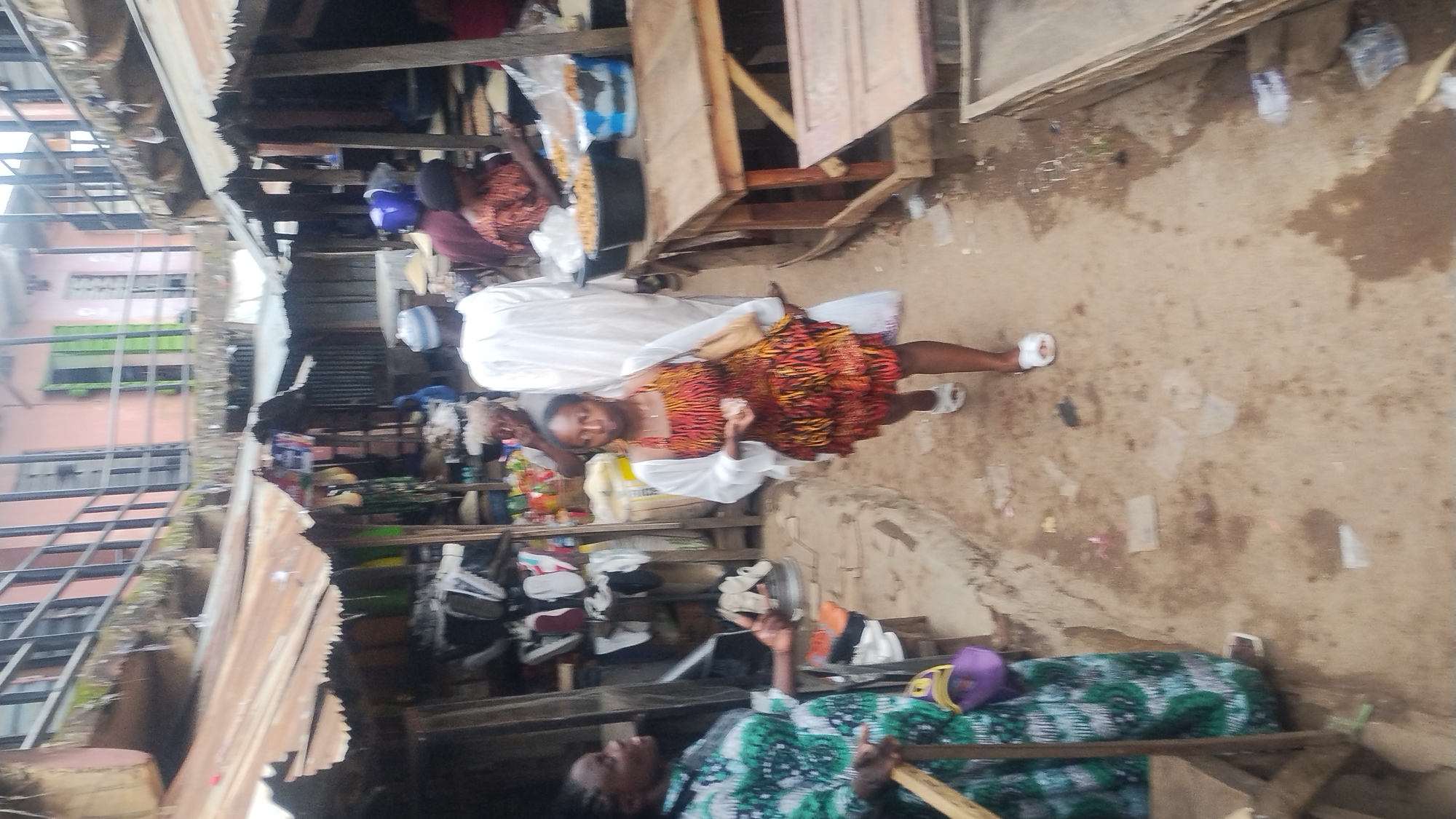 The image shows an outdoor market scene. A young woman wearing a colorful, patterned dress and a white cardigan walks towards the viewer. She is carrying a beige handbag on her right shoulder and a white plastic bag in her left hand. To the left of the woman, a woman in a green and white patterned dress stands facing her, seemingly engaged in a conversation. The background reveals market stalls with various goods, including shoes on shelves, bags of produce, and buckets of food items. Several other individuals are visible in the market, some in traditional clothing. The ground is dusty and appears to be uneven, with some debris scattered around. The structures of the market appear to be made of wood and corrugated metal.