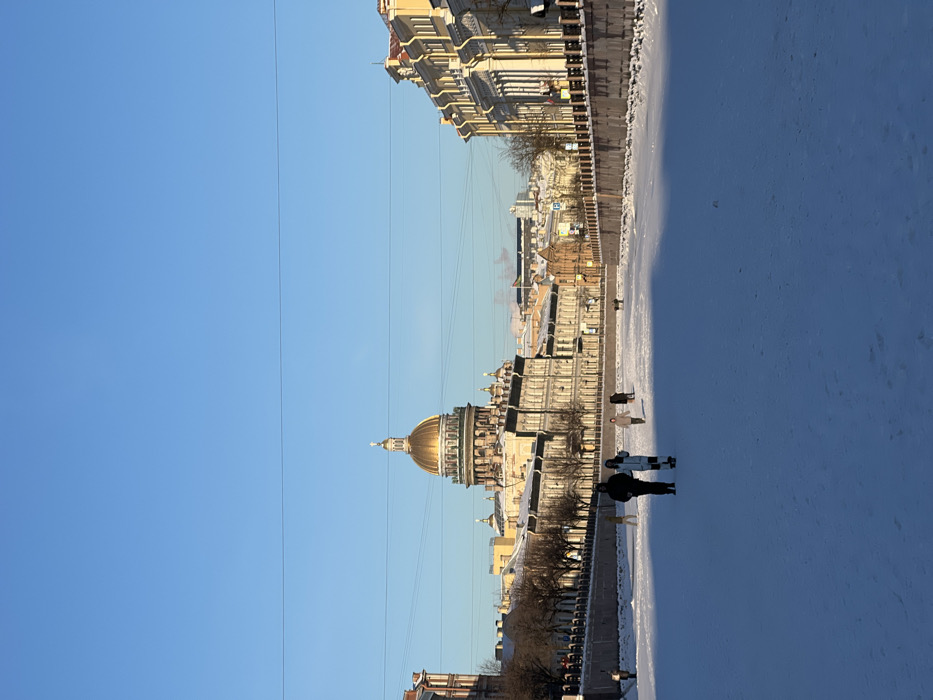 This image depicts a winter scene in Saint Petersburg, Russia, looking north-northwest along a frozen canal, likely the Moika River, towards Saint Isaac's Cathedral in the distant background. The cathedral is identifiable by its prominent golden dome, smaller cupola, and a cross at its apex. The sky is clear and bright blue, suggesting daytime.
Sunlight illuminates the multi-story historic buildings lining the right bank of the frozen waterway, revealing classical architectural elements, including visible columns. Buildings along the left bank are partially shaded, with bare trees bordering the stone embankment. The canal itself is entirely frozen and covered with a uniform layer of snow, creating a wide, white surface.
Multiple human figures are present on the frozen canal. In the mid-foreground, two individuals are walking from left to right. The lead person is dressed in dark winter clothing. The second person, wearing light-colored winter attire, is positioned slightly behind and to the right of the first, and appears to be interacting with or manipulating a small, dark object on the ice. At least five additional human figures are discernible further in the background, also walking on the frozen surface. Thin, dark power lines are visible stretching horizontally across the upper third of the sky.