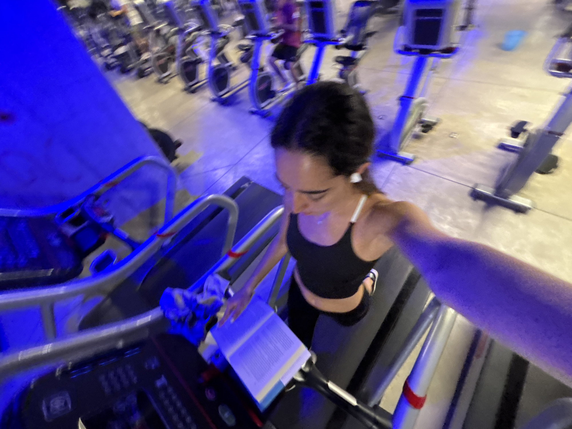 A woman in a black crop top and black leggings walks on a treadmill while reading a book at a gym. The gym appears to be lit with blue and yellow lighting, and there are several stationary bikes in the background.