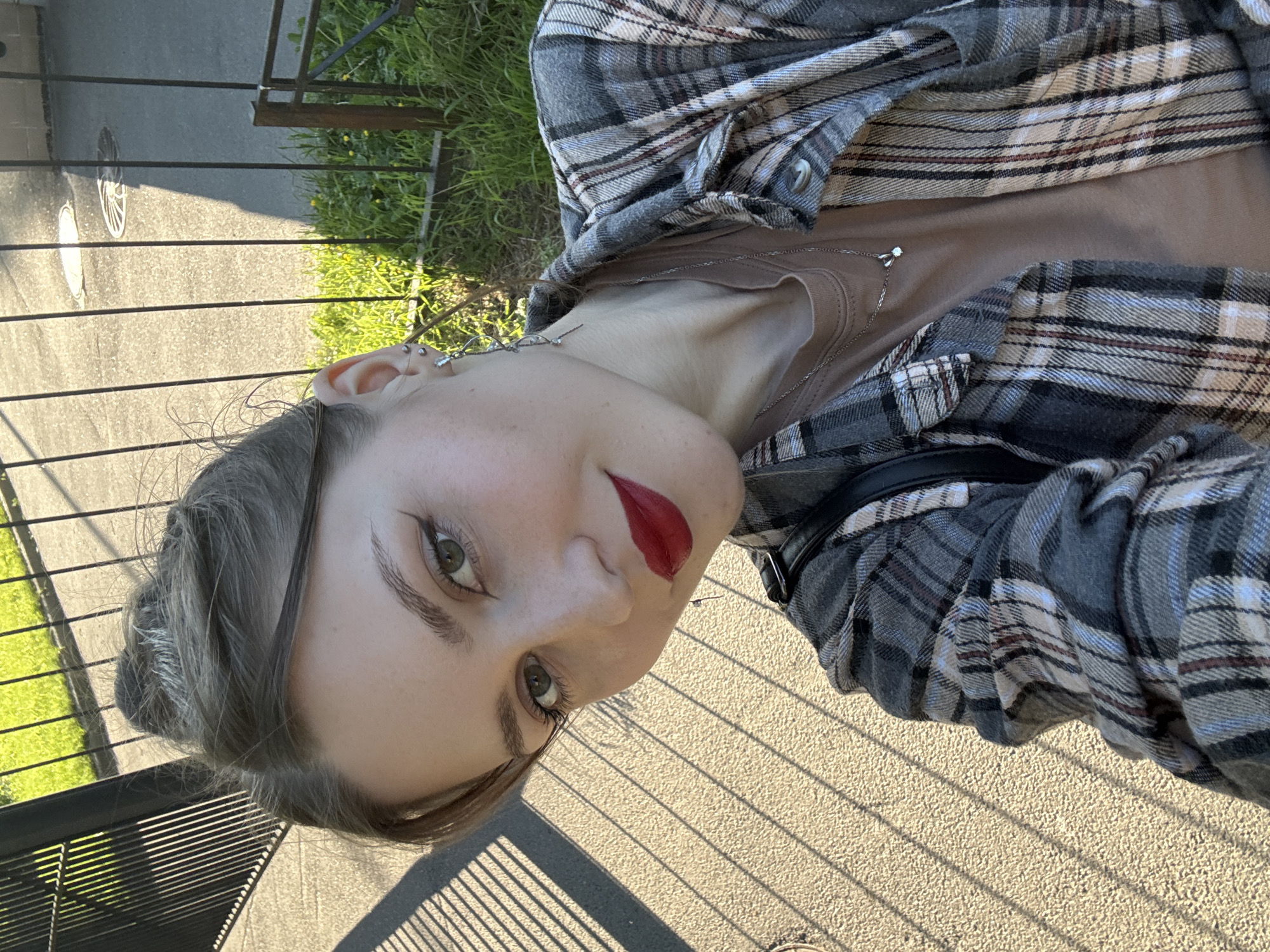 A young woman, appearing to be in her twenties, is captured in a close-up, upward-angled selfie, looking directly at the camera with a subtle smile. Her hair, a light brown or ash tone, is pulled back into a messy bun at the crown of her head. She has green eyes accentuated with black winged eyeliner and mascara, and well-defined eyebrows. Her lips are covered in bold red lipstick. She is wearing a light brown crew-neck t-shirt layered under an open flannel shirt with a plaid pattern featuring shades of gray, white, black, and thin maroon stripes. A thin silver-colored necklace with a small, faceted pendant is visible around her neck. Her left ear displays multiple piercings, including a helix piercing and an elaborate, silver-toned earring resembling abstract foliage or branches. A black strap, likely from a bag, is visible over her right shoulder.
The scene is outdoors, in what appears to be the city of Saint Petersburg, Russia, under strong natural lighting, likely direct sunlight, which casts long, distinct parallel shadows from a vertical barred fence onto the light-colored, textured pavement in the foreground and to the left. In the background, behind a darker metal fence, a strip of green grass with scattered small yellow flowers is visible, beyond which lies a darker asphalt surface with two manhole covers and a light-colored building wall. The lighting suggests either early morning or late afternoon.
