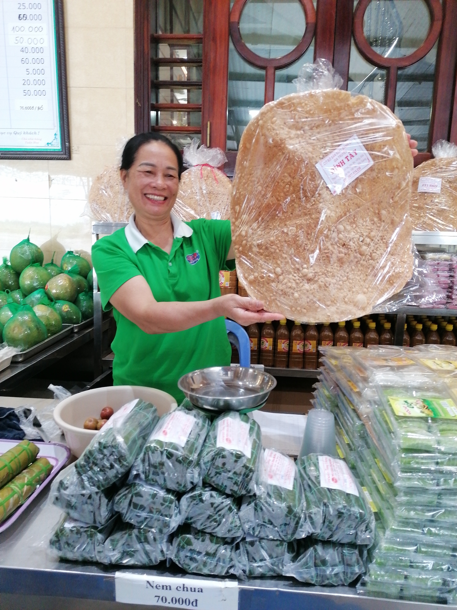 Inside a shop in Chu Văn An Ward, Vietnam, a smiling Asian woman stands behind a counter laden with various food items. She holds a large, round, flatbread-like product in a plastic wrapper. Behind her are shelves stocked with packaged food and bottled sauces. To her left, a stack of green-skinned fruits sits on a tray. In front of her, the counter displays neatly packaged bundles, a bowl of small round objects, and stacks of clear-wrapped food items. A sign on the counter reads "Nem chua 70.000d". Above the woman and to the left is a framed price list. The shop's background reveals a window with circular designs.