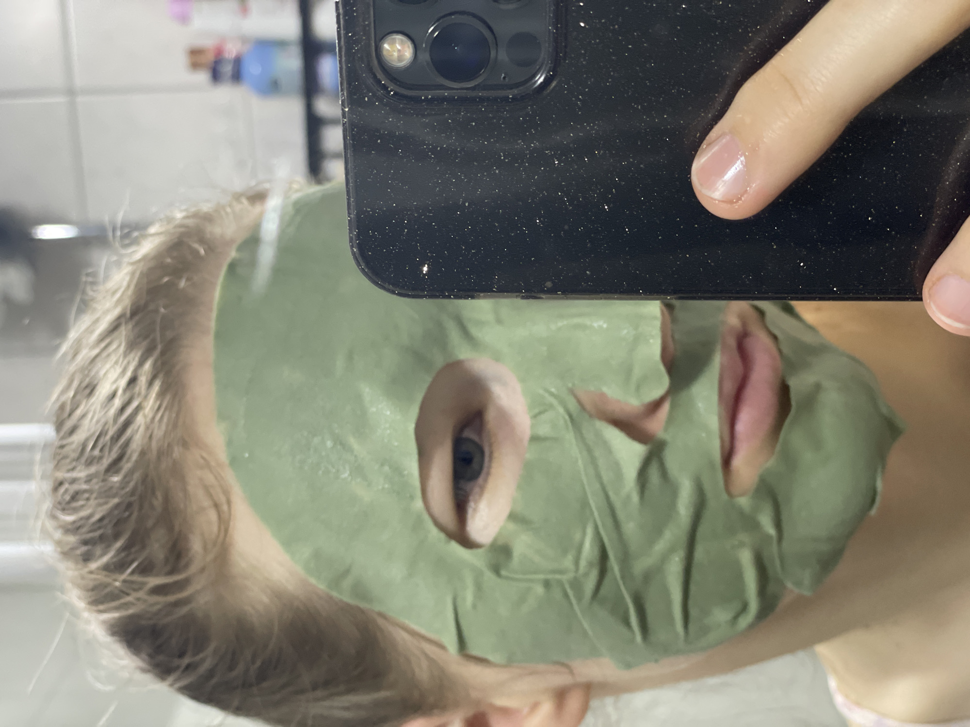 A young Caucasian person with fair skin and blonde hair is taking a selfie in a bathroom. They are wearing a green sheet mask that covers most of their face, revealing one eye and their mouth. The person is holding a black smartphone in their right hand, with their index finger resting on the screen. The background is blurred but appears to be a bathroom with tiled walls and shelves containing various bottles and products. The lighting is bright, suggesting the presence of overhead lights or natural light from a window. The image is focused on the person's face and the smartphone. There is no indication of the city of Nidda, Germany in the image.