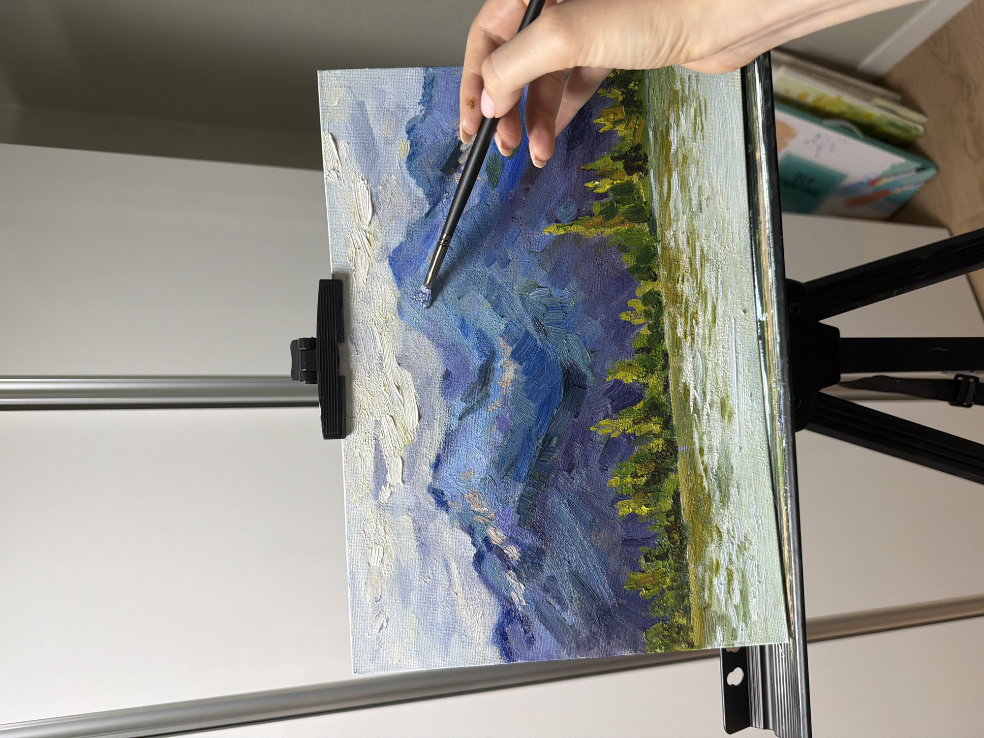 An artist's hand holding a paintbrush is visible in the foreground, about to add a touch of paint to a landscape oil painting. The painting, which is displayed on a black easel, depicts a mountain range with a body of water in the foreground and trees lining the shore. The sky above the mountains is filled with clouds. The background of the image shows a portion of a white wall, suggesting an indoor setting, possibly a studio or a room in a residential building in Dongducheon-si, South Korea.