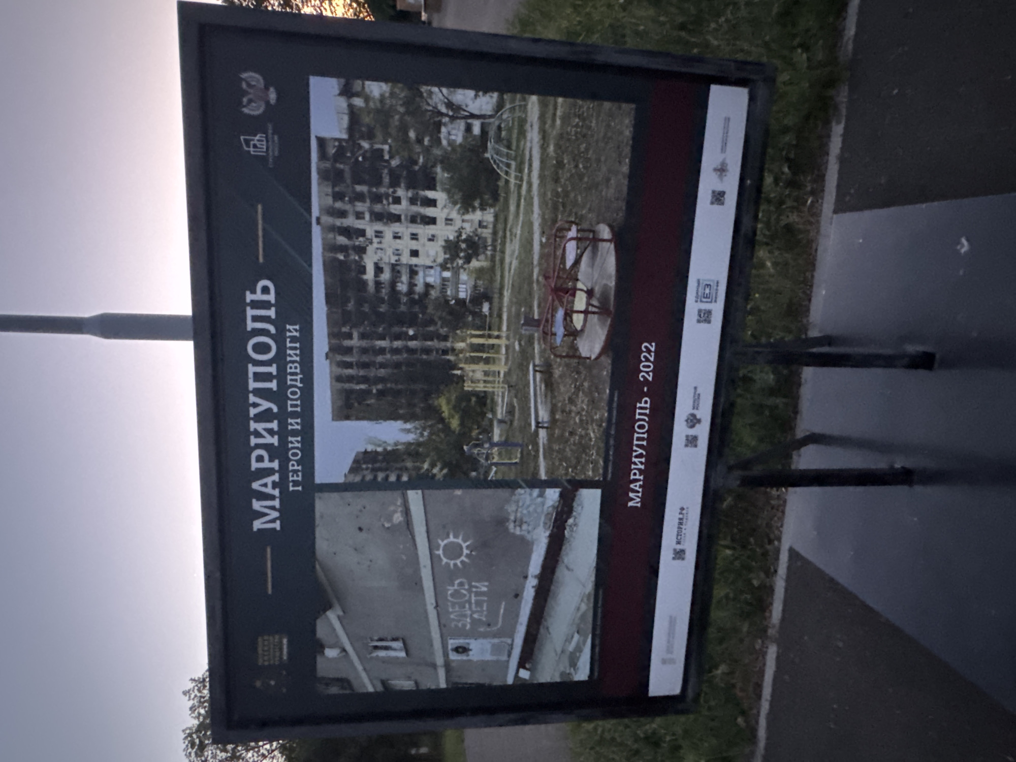The image shows an outdoor exhibition display in the city of Mariupol, Ukraine. The display features two photographs side-by-side, presented on a dark background with text and logos. The overall tone is somber, reflecting the subject matter.
The left photograph shows a damaged apartment building with visible bullet holes and rubble. A white message spray-painted on the wall reads "ЗДЕСЬ ДЕТИ" (Here are children), with a drawing of a sun next to it. This message is positioned near a doorway. The building appears to be weathered and neglected.
The right photograph depicts a playground in front of a severely damaged apartment building. The building is extensively damaged, with many windows broken or missing, and parts of the facade are charred or destroyed. The playground equipment, including a merry-go-round, swings, and climbing structures, is visible amidst the debris and overgrown vegetation. The scene suggests a former residential area impacted by conflict.
The text at the top of the display reads "МАРИУПОЛЬ" (Mariupol) and below it, "ГЕРОИ И ПОДВИГИ" (Heroes and Feats). The bottom of the display includes the text "МАРИУПОЛЬ - 2022" (Mariupol - 2022) and various logos, including one that appears to be the Russian coat of arms. The display is mounted on a dark metal stand and is situated outdoors, likely in a public space, with a paved area in the foreground and grass and trees in the background. The sky above is overcast with hints of sunset. The overall impression is one of documenting destruction and loss in Mariupol during 2022.