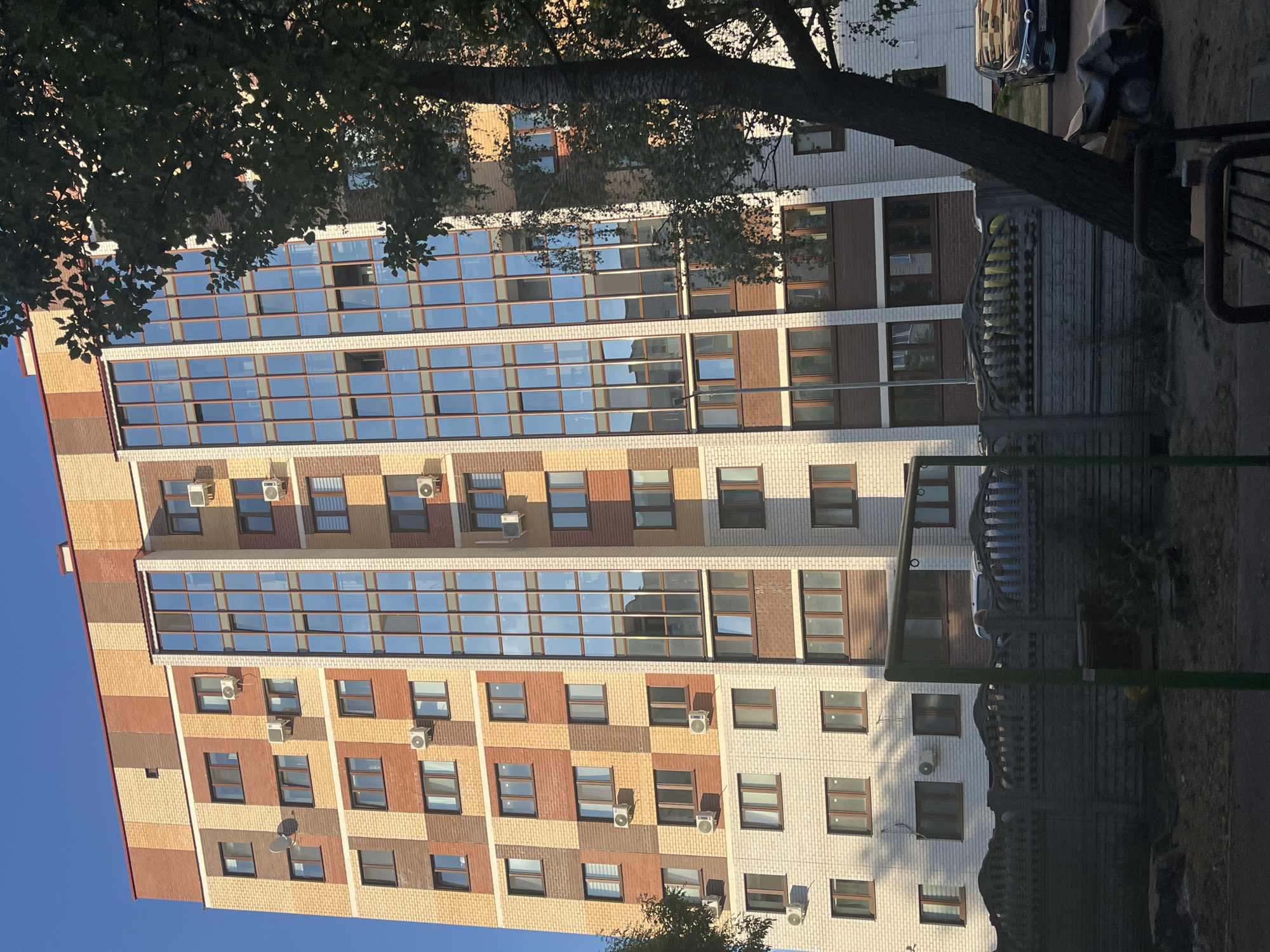 A multi-story residential building with a facade of varied colored bricks and large glass windows is depicted. The building features numerous air conditioning units mounted on the exterior walls. In the foreground, a concrete fence partially obscures a street with parked cars and trees. The sky is clear and blue, suggesting daylight.